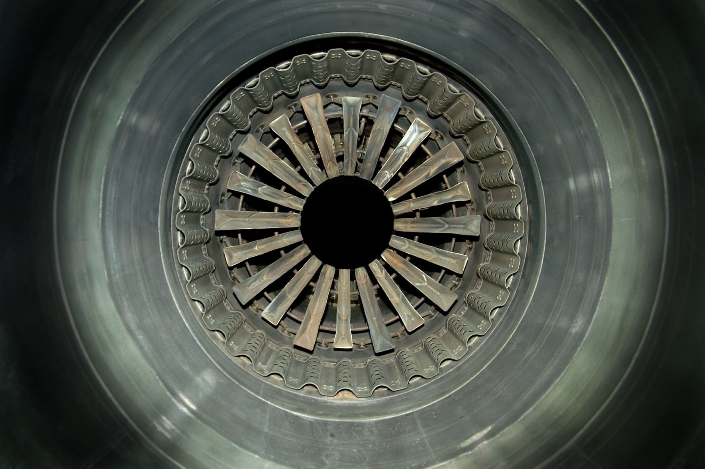 Frontal view of a jet engine intake, showing concentric circles of metallic blades and ducts radiating from a central black aperture. The photograph captures the geometric precision and industrial symmetry of the engine's interior in cool gray and silver tones.