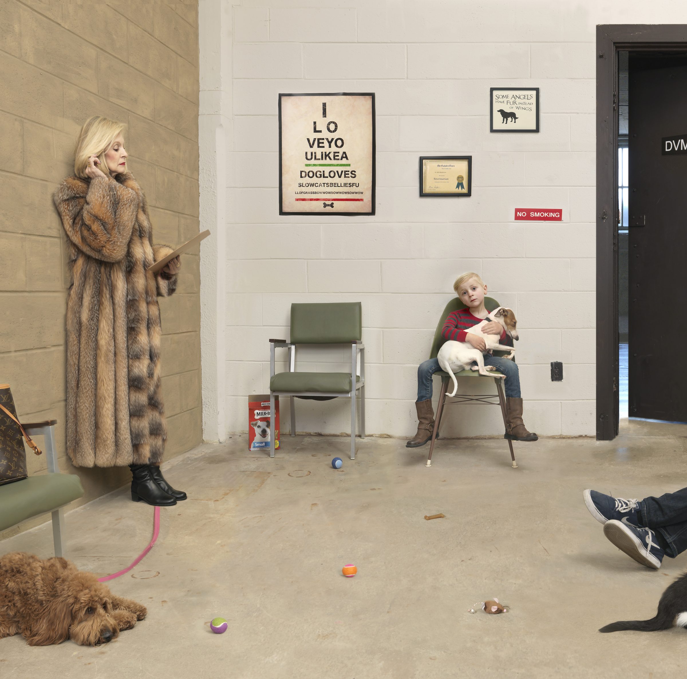 A woman in a fur coat stands beside a man in dark shoes with legs stretched out on the floor, while a young boy sits holding a small dog in a green chair. Dogs and toys scatter across a concrete floor in a clinical waiting room with white brick walls, framed dog-themed posters, and a "No Smoking" sign.