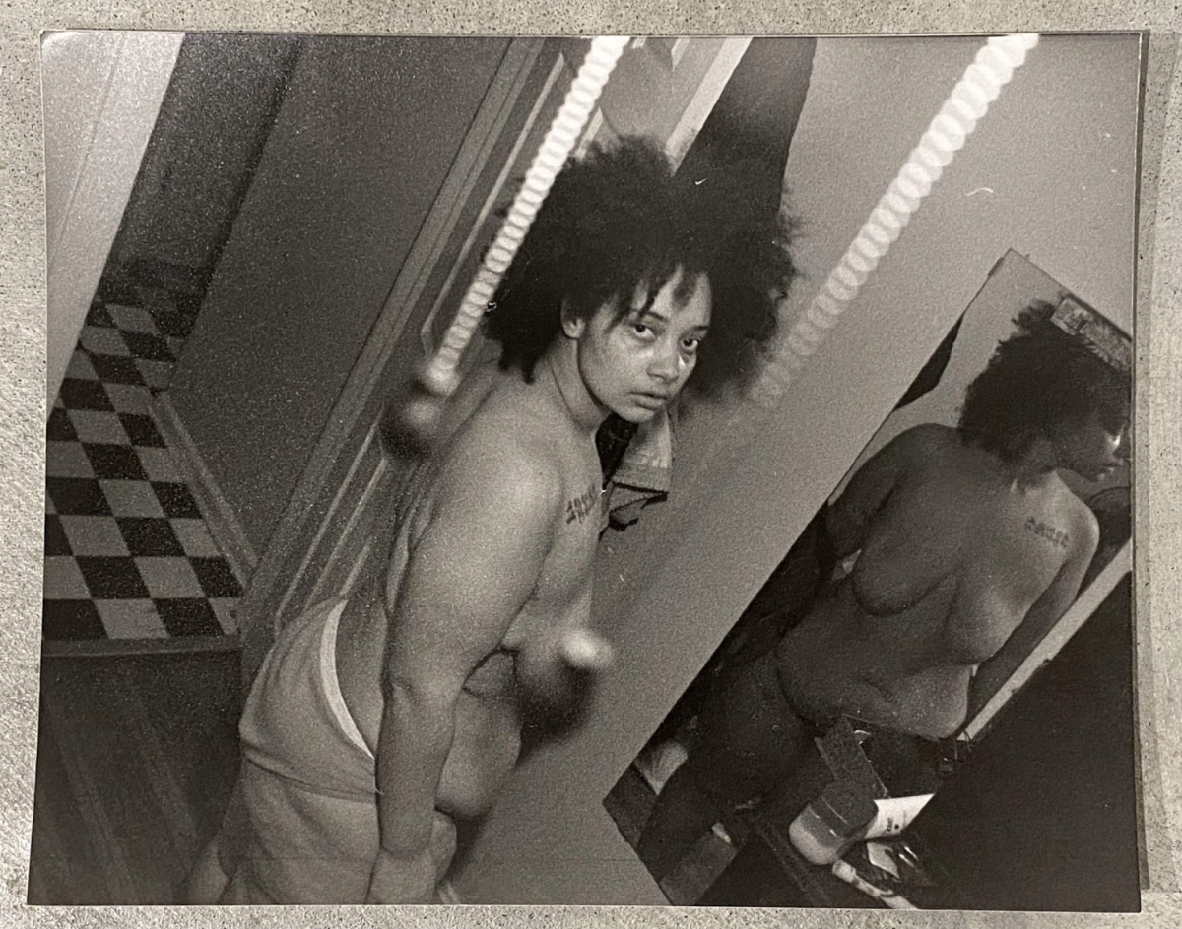 Black and white photograph showing two shirtless figures in an interior space with fluorescent ceiling lights and patterned flooring. The figure on the left, with voluminous dark hair, gazes toward the camera with a contemplative expression, while a second figure is visible on the right side of the frame. The composition captures an intimate domestic moment rendered in stark, high-contrast tones characteristic of silver gelatin printing.