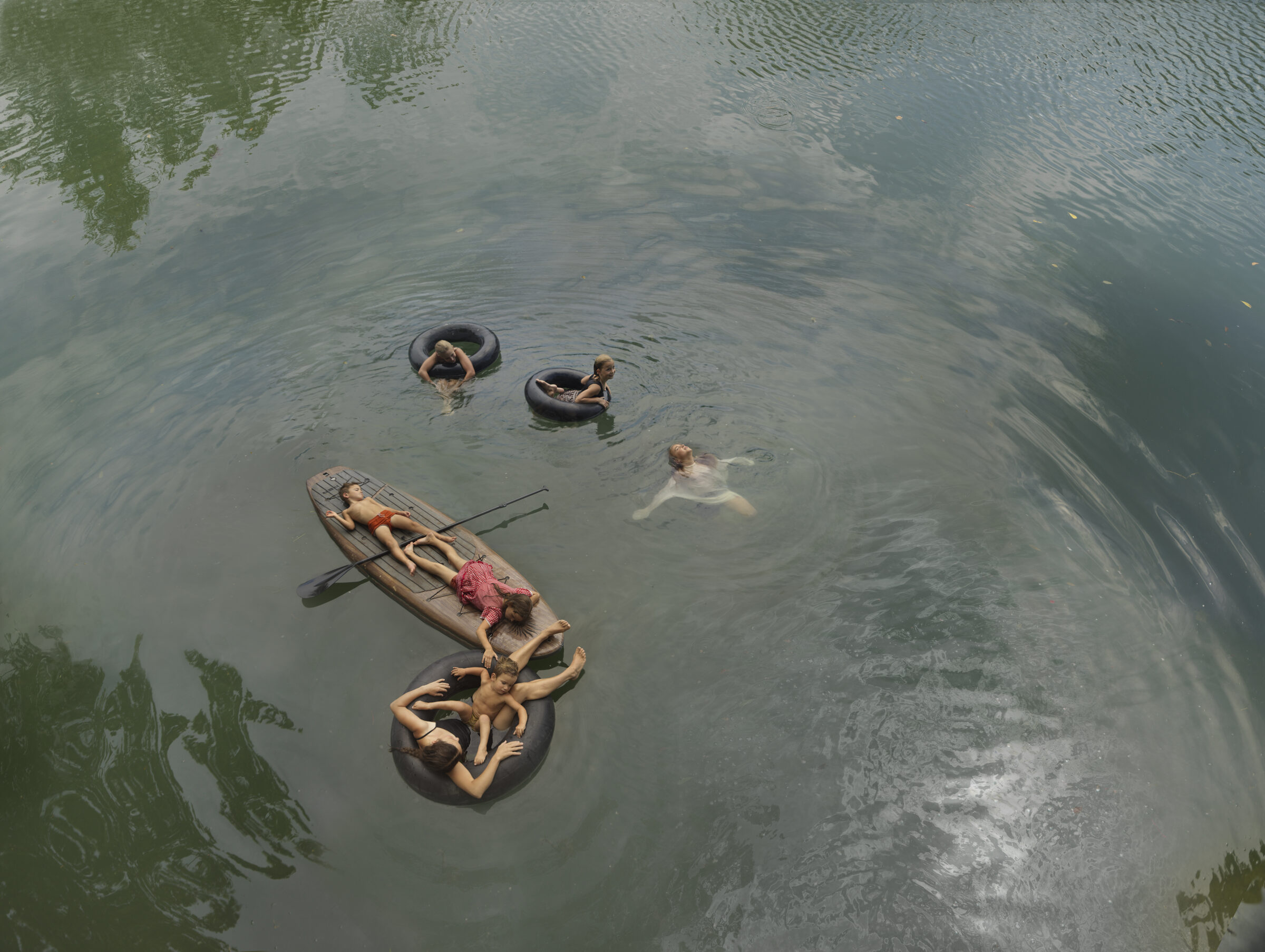 Aerial view of a murky river where multiple figures are scattered across the water—some floating on inner tubes, others swimming or reclining on a small boat with oars. Vegetation is visible along the banks, and the water's surface reflects muted greens and grays in a languid summer scene.