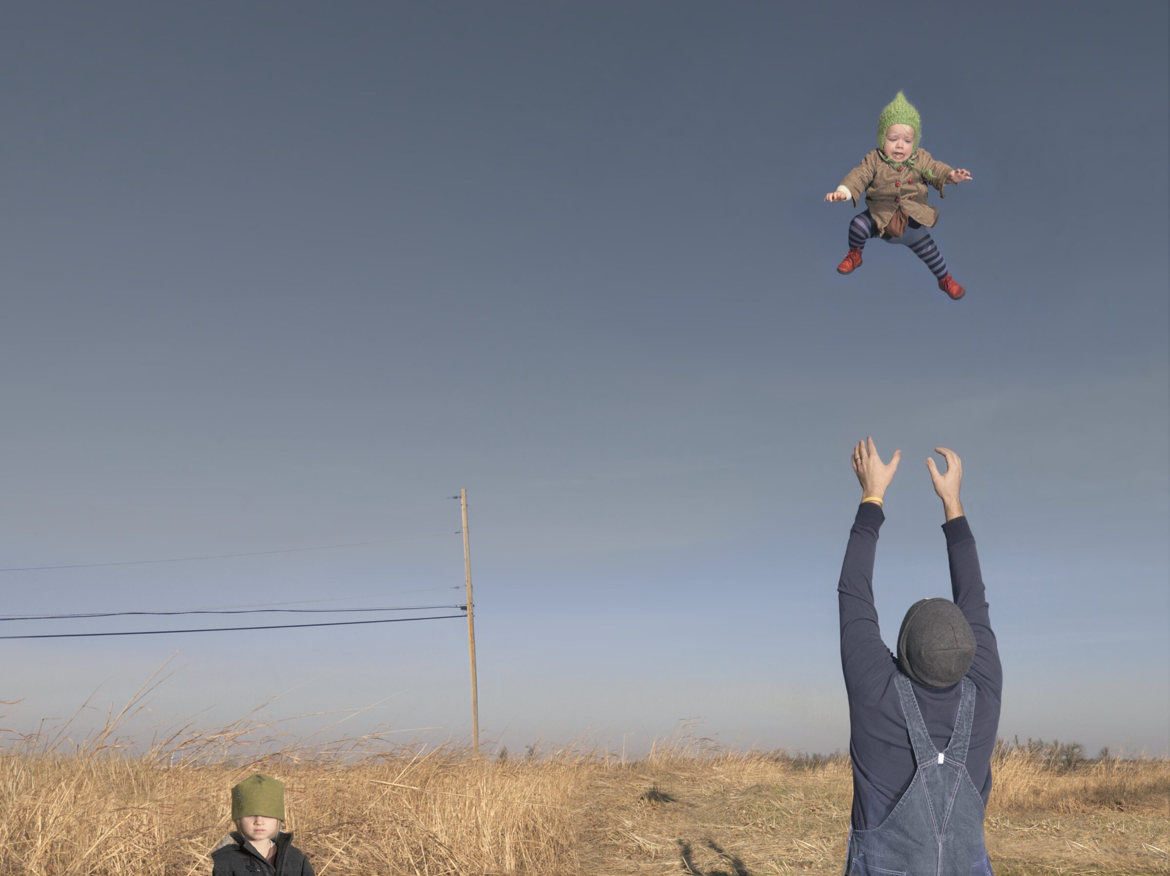 An adult wearing a dark jacket and green hat tosses a small child in a green knit hat high into a clear blue sky above a vast, dry grass field. Another child in a green hat stands in the foreground as the adult's arms reach upward to catch the airborne child, with a utility pole visible on the horizon.