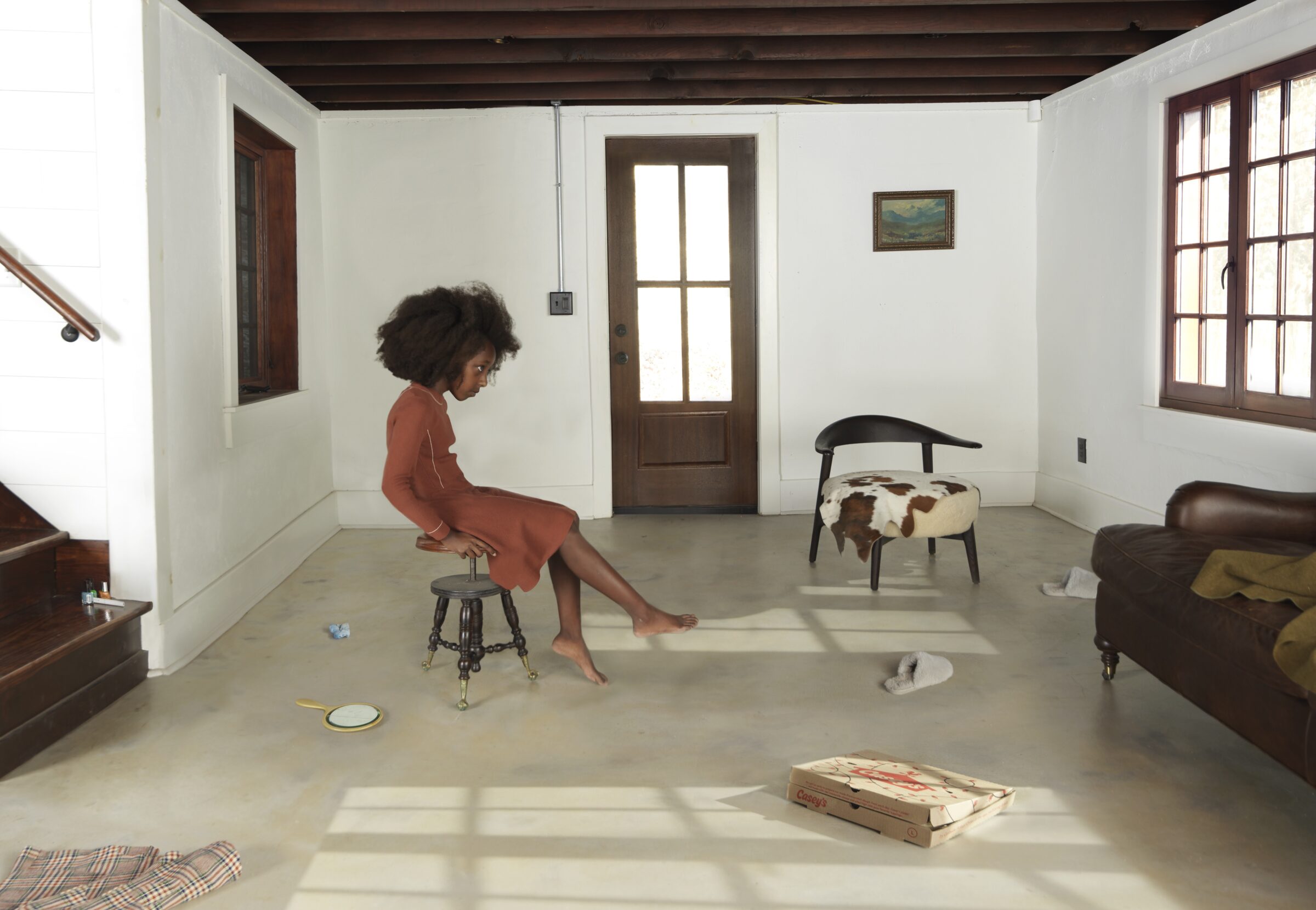 A woman in a rust-colored dress sits on a wooden stool in a sparsely furnished, sunlit room with concrete flooring, white walls, and dark wooden architectural elements. The minimalist interior contains scattered objects including chairs, a couch, pizza boxes, and dishes, creating an ambiguous domestic scene composed with careful attention to light and shadow.