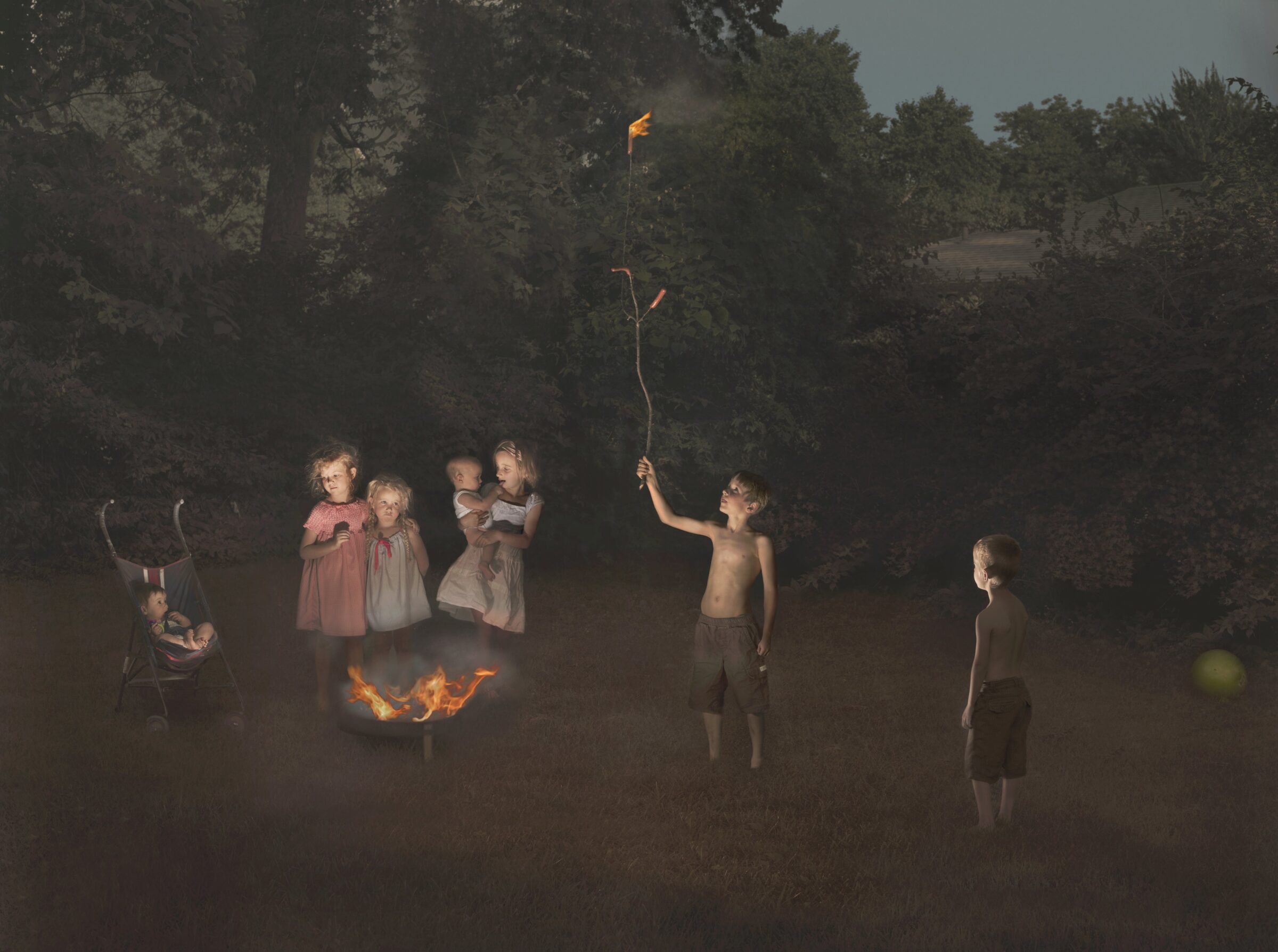 Children gathered in a misty forest clearing around a campfire, with one child holding aloft a flaming torch against dark trees. The scene is rendered in muted, atmospheric tones with dramatic lighting that isolates the figures from the shadowy woodland background.