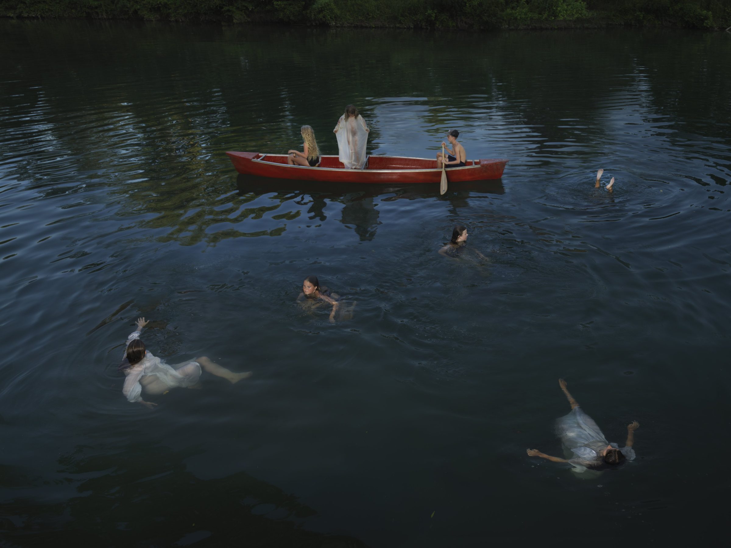 Multiple figures in white clothing swim in dark water around a red rowboat, with two people seated in the boat and dense green foliage visible along the shoreline in the background.