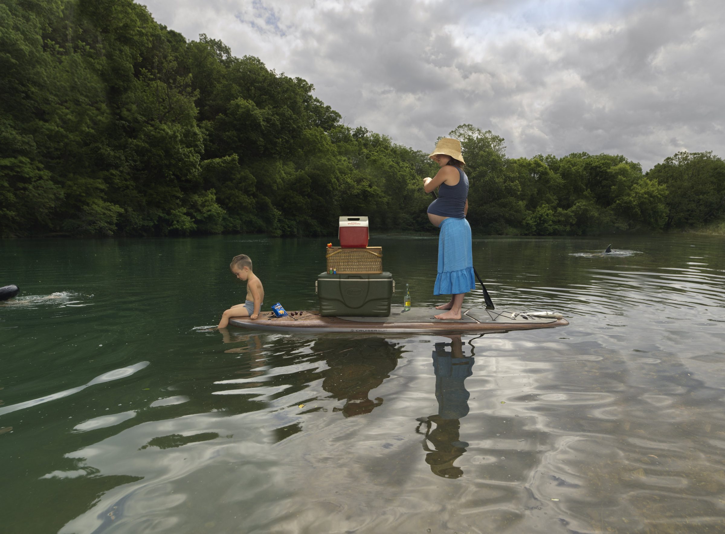 A woman in a straw hat and blue skirt stands on a paddleboard in shallow river water, holding a paddle and gazing outward. A young child sits in the water beside her, while various domestic items—a cooler, picnic basket, and other supplies—are stacked on the board behind her. Dense green forest lines the riverbank under an overcast sky.
