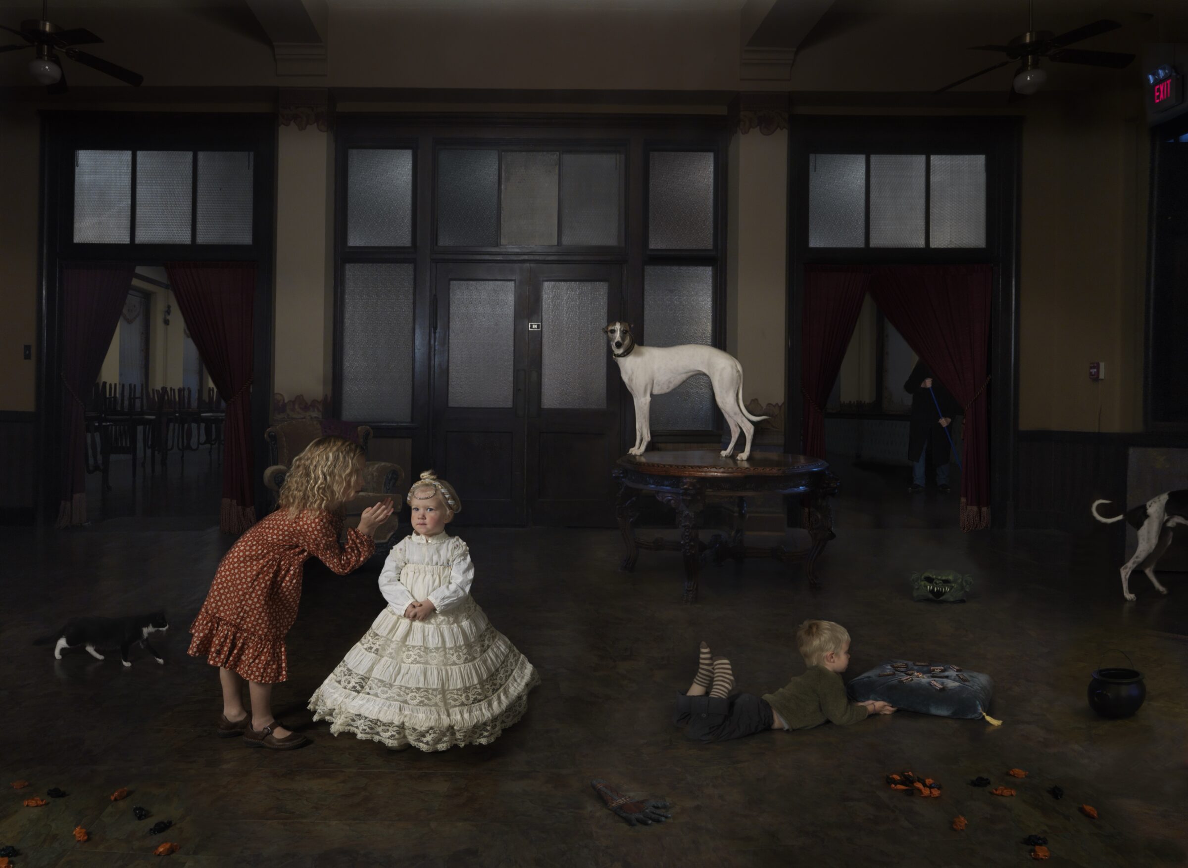 A meticulously composed interior scene depicts children in period dress interacting with multiple dogs in a modernist gallery space with dark-framed glass doors and frosted panels. A child in an ornate white dress stands center while another in a rust-colored coat bends toward her; additional children play on the polished floor as white and spotted dogs occupy various positions throughout the room, creating a surreal, theatrical tableau.