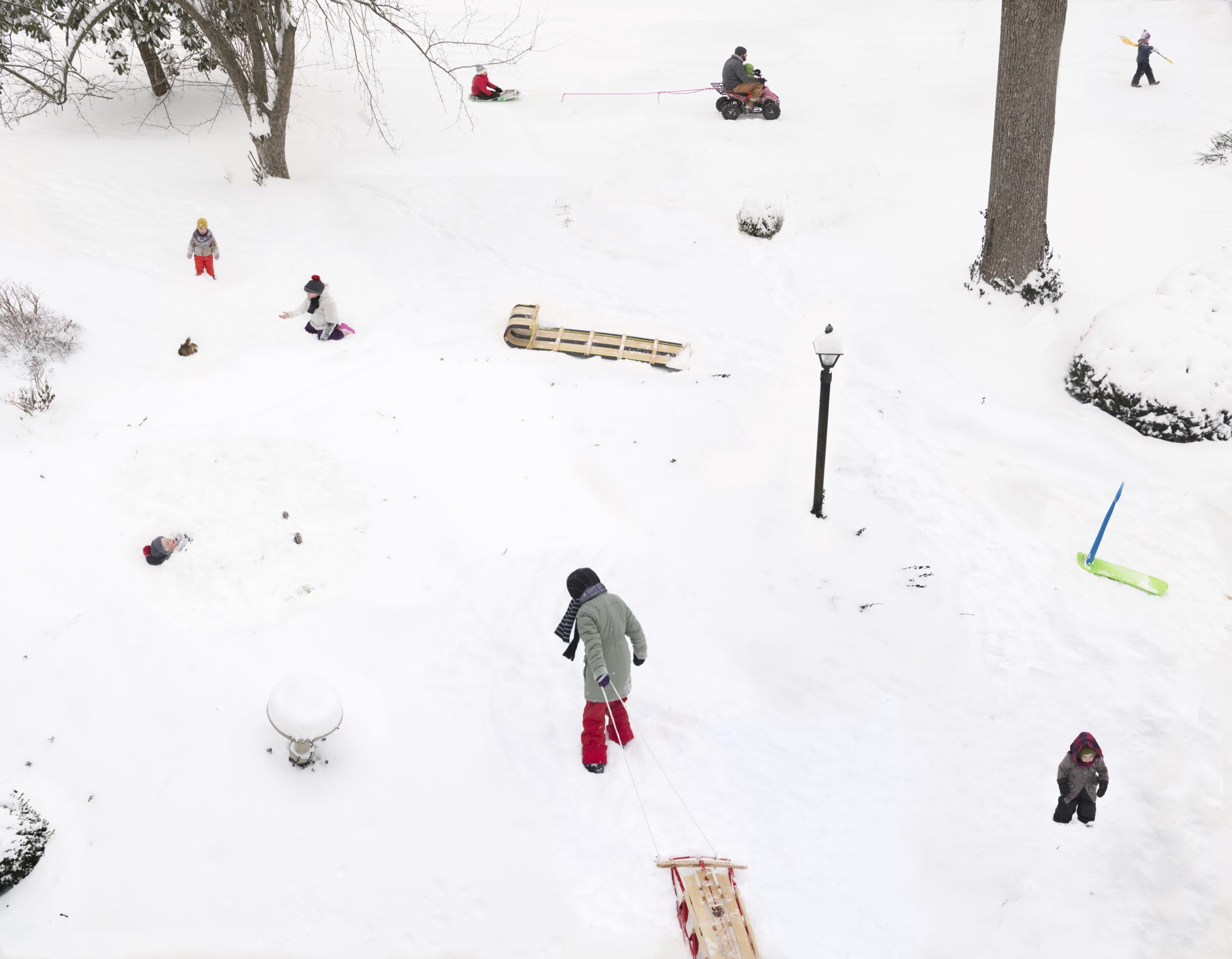 Aerial view of a snowy hillside with multiple people engaged in winter activities scattered across the landscape, including sledding, snowmobiling, and walking. The composition captures the scene from above, with bare trees, evergreen shrubs, and various wooden sleds distributed throughout the white expanse, creating a lively tableau of winter recreation.