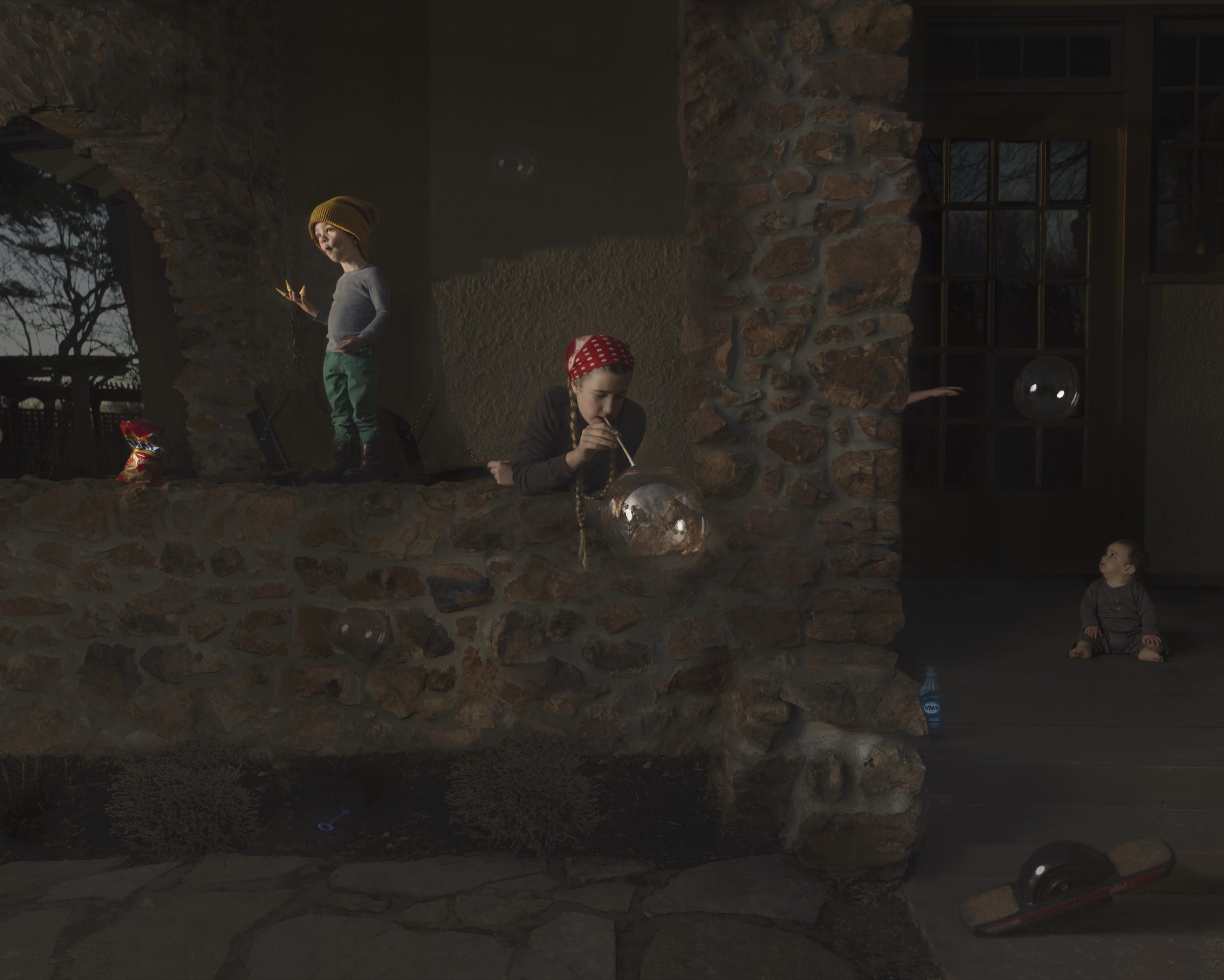 Three children play in a shadowed stone courtyard lit by warm, directional sunlight. One child in a yellow hat stands against the wall on the left, another in a red bandana blows bubbles in the center, and a third sits in darkness on the right. The weathered stone walls and sparse setting create a contemplative atmosphere punctuated by the ephemeral quality of soap bubbles.