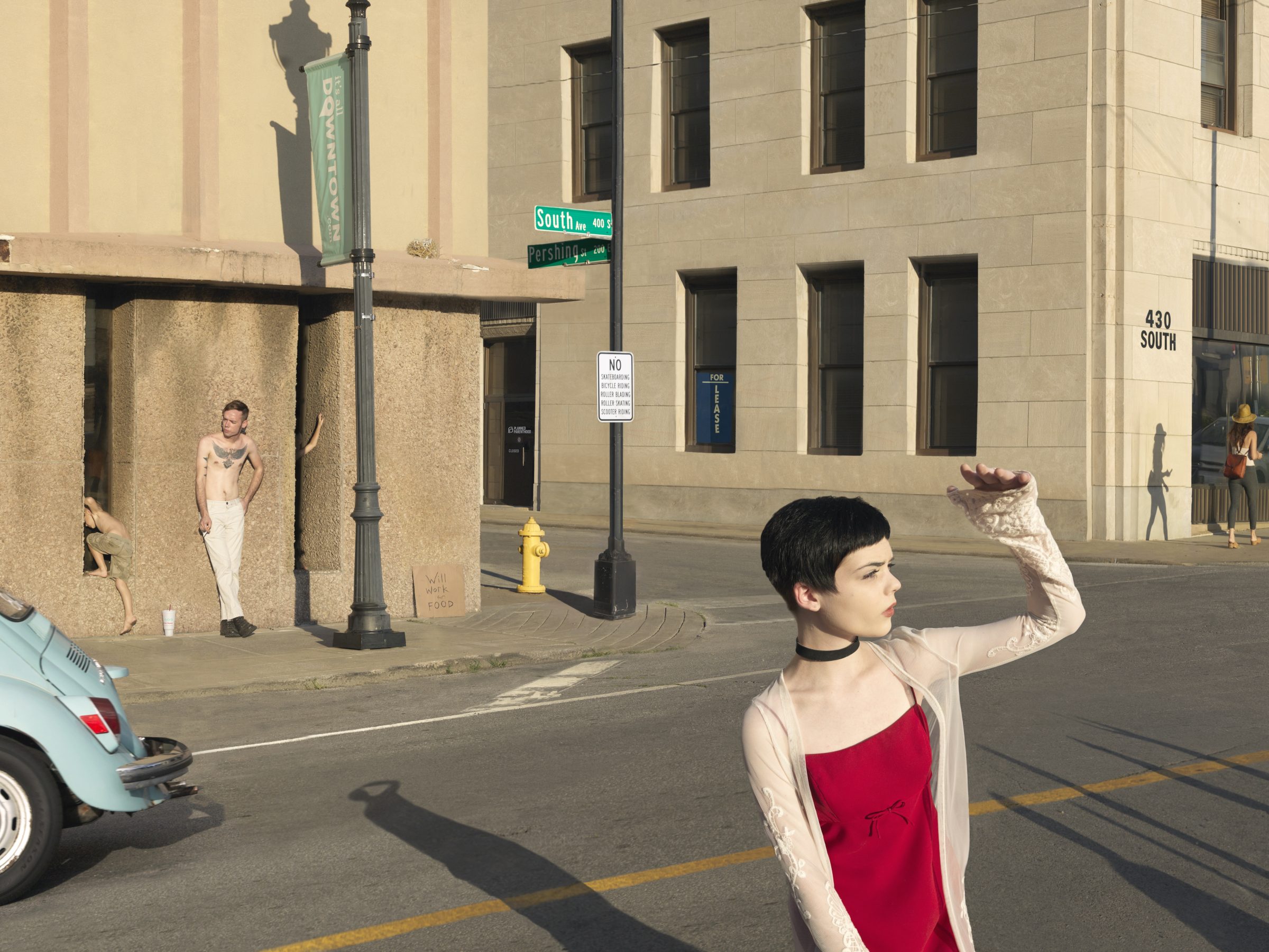 A woman in a red dress and beige cardigan stands in an urban street intersection, shielding her eyes while looking upward. Behind her, figures are visible near a concrete structure at a street corner marked by blue "South" and "Pershing" street signs, with a classical stone building facade and parked car visible in the composed scene.