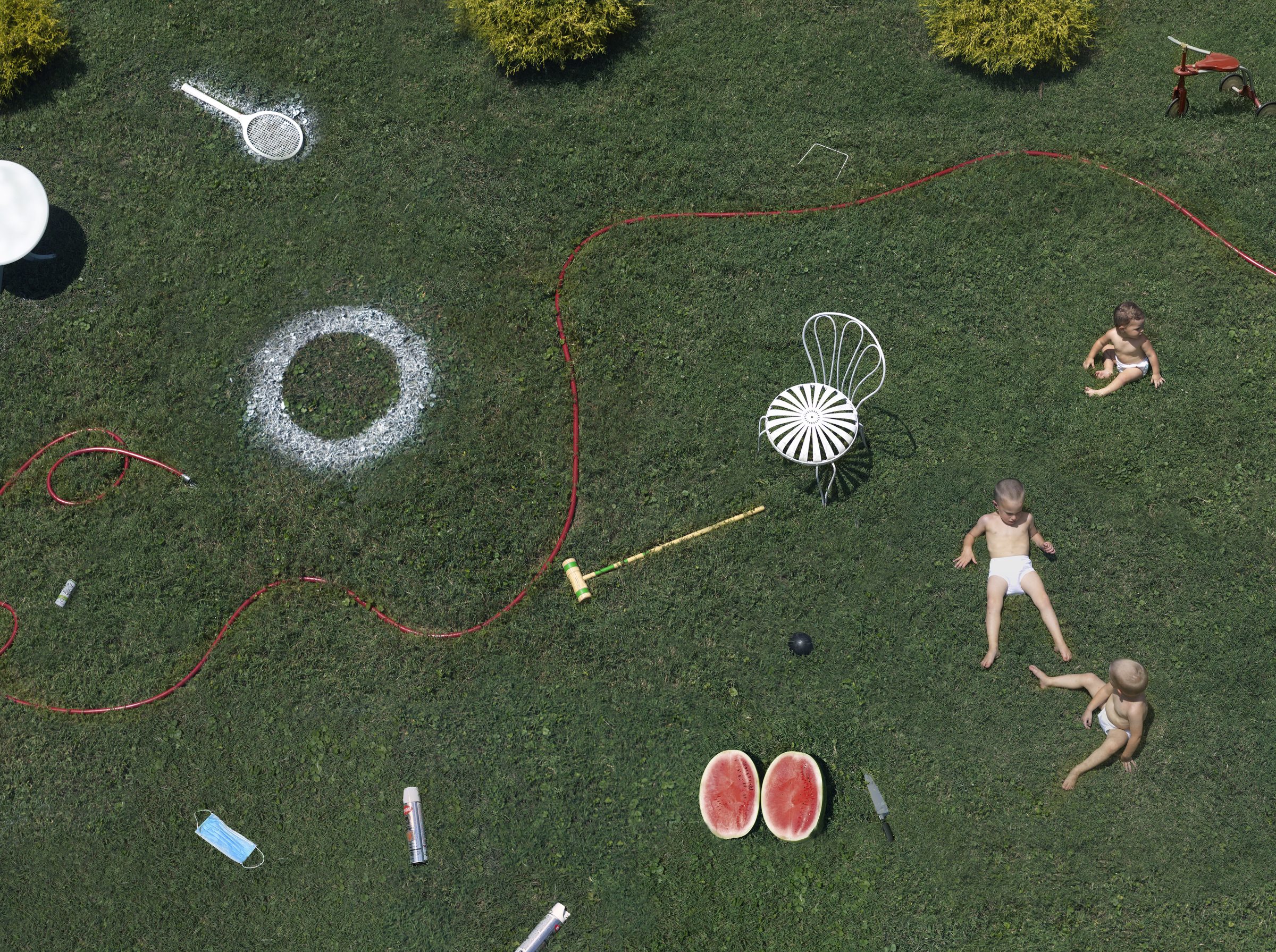 Aerial view of a lush green lawn scattered with summertime objects and three shirtless children in motion. White garden furniture, a red hose, chalk circles, watermelon halves, various toys, and garden tools are arranged across the grass in a vibrant, playful composition.