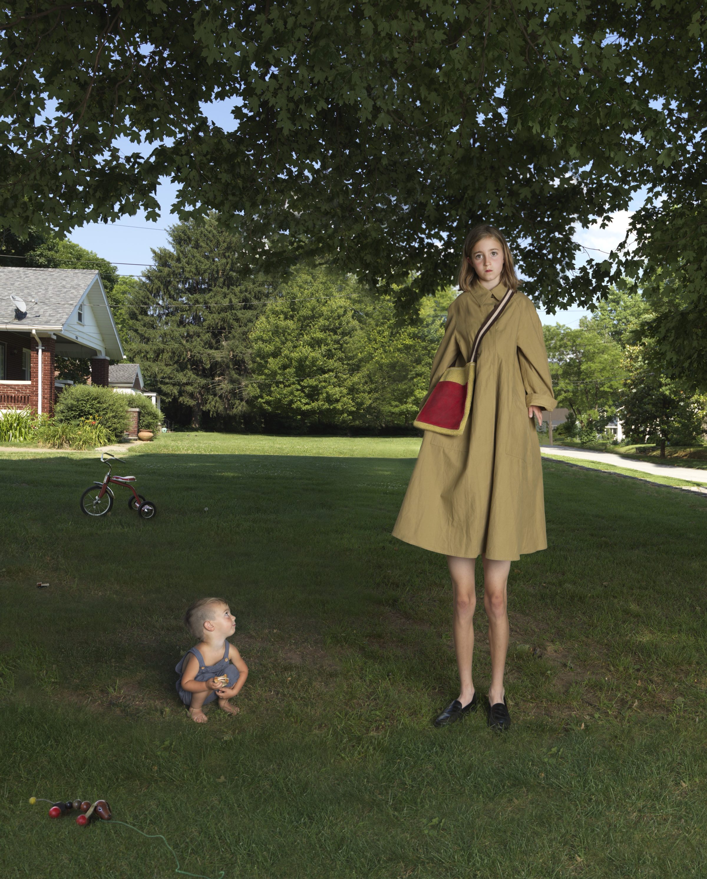 A young woman in a tan coat with a burgundy bag stands in a suburban lawn, her proportions digitally altered to an unnaturally tall height. She gazes downward at a toddler crouching nearby, while toys, a tricycle, and a brick house populate the verdant yard behind them under a canopy of leafy trees.