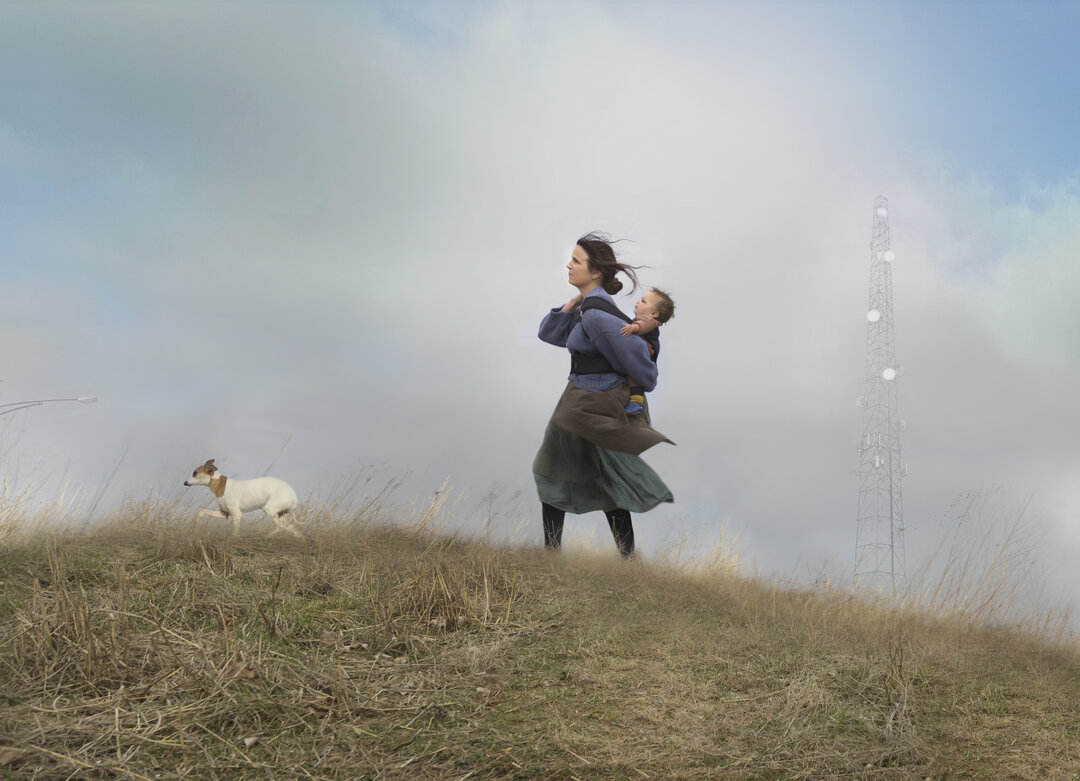 A woman holds a small child on a barren hilltop dotted with dry grass and vegetation, while a small dog stands nearby. A tall communication tower rises through the misty background on the right side of the composition, creating a stark contrast between the intimate human figures in the foreground and the industrial structure beyond.