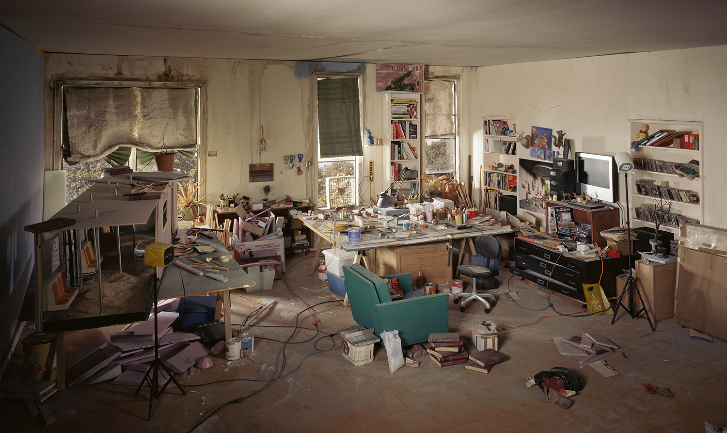 A meticulously detailed interior space cluttered with art supplies, books, and personal objects scattered across a concrete floor. Sunlight streams through windows and glass doors, illuminating shelves lined with books and artwork, a turquoise armchair, wooden furniture, and various materials in warm, naturalistic tones that contrast with the industrial setting.