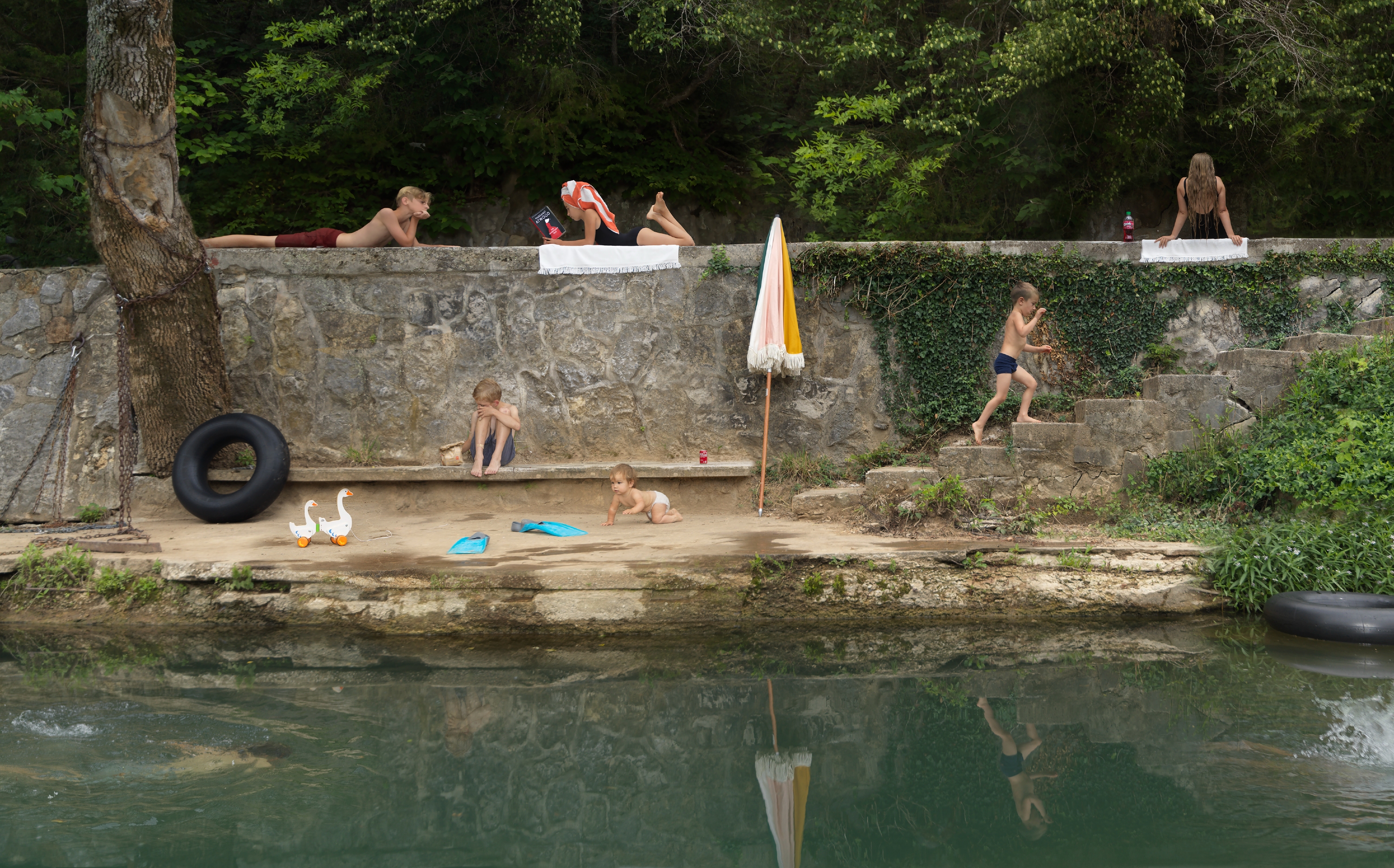 A leisurely riverside scene where multiple children and adults occupy a tiered stone embankment in various states of summer play and relaxation. Some figures recline on the upper ledge, others wade or sit along the sandy beach below, while a pair of white geese, beach toys, and an umbrella populate the foreground. Dense green foliage frames the scene, and the still water below mirrors the activity above.