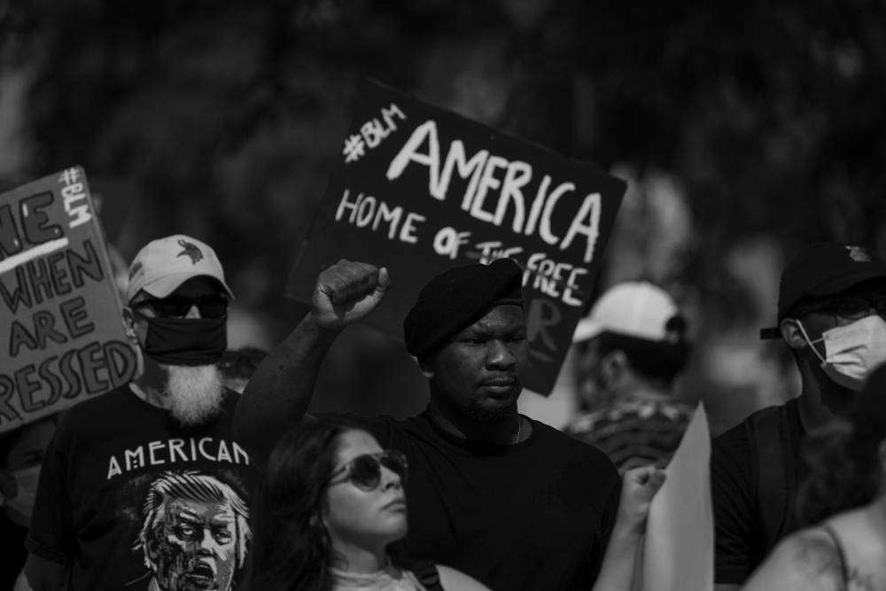 Black and white photograph of a protest crowd holding signs, including one reading "AMERICA HOME OF THE FREE." Participants wear masks and caps, with raised fists visible among the group, capturing a moment of civil demonstration and activism.