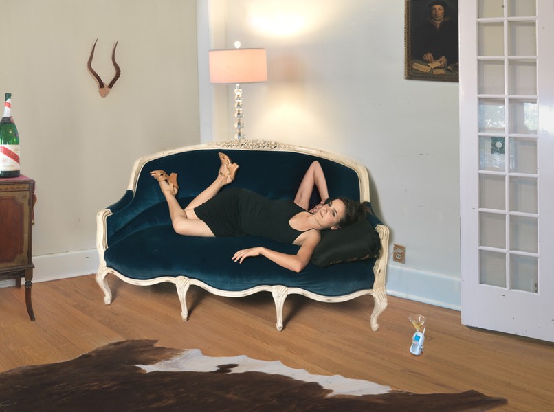 A woman in a black dress reclines on an ornate cream-and-blue Rococo-style sofa in a contemporary interior space. The staged domestic scene includes period furniture, decorative objects, and natural lighting, with a cowhide rug visible on the wooden floor.