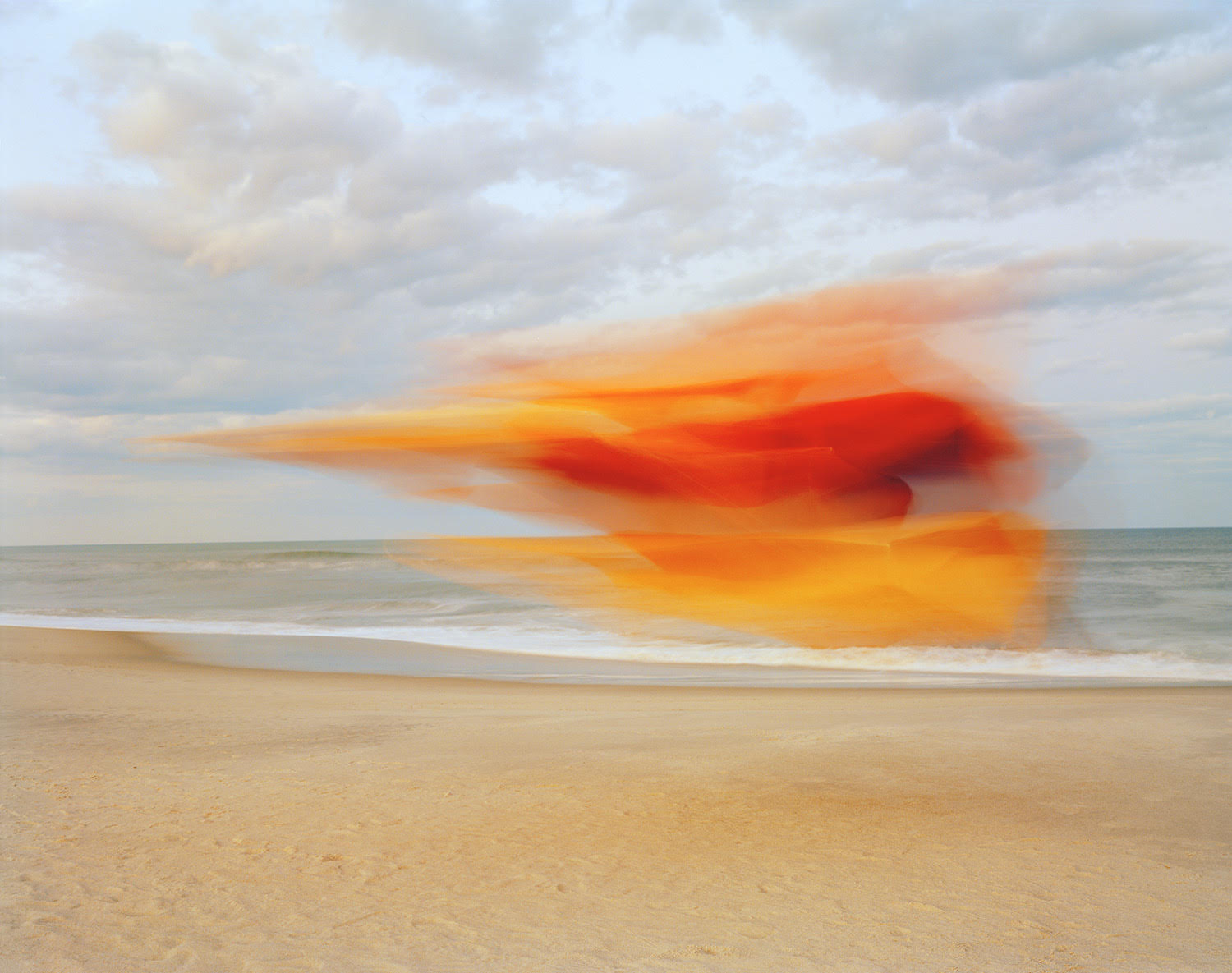 A vibrant abstract blur of red and orange streaks sweeps horizontally across a serene beach landscape, creating a sense of motion against the still horizon where sand meets calm ocean waters under an overcast sky.