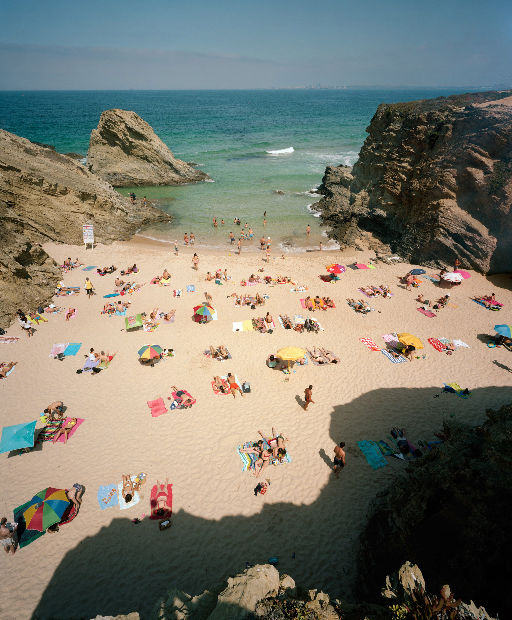Aerial view of a crowded sandy beach nestled between dramatic rocky outcroppings, with colorful umbrellas and beachgoers scattered across the shore, turquoise water and breaking waves visible beyond.