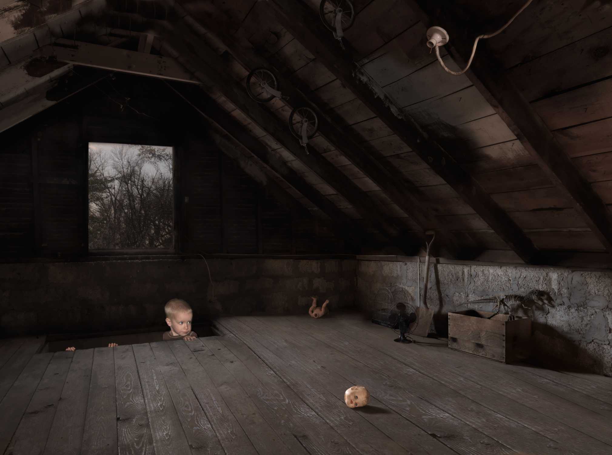 A young child peers over wooden floorboards in a sparse, dimly lit attic space, with bare wooden beams overhead and a window showing bare winter trees. A doll's head rests on the floor in the foreground, while another doll and various objects are scattered throughout the shadowy interior.