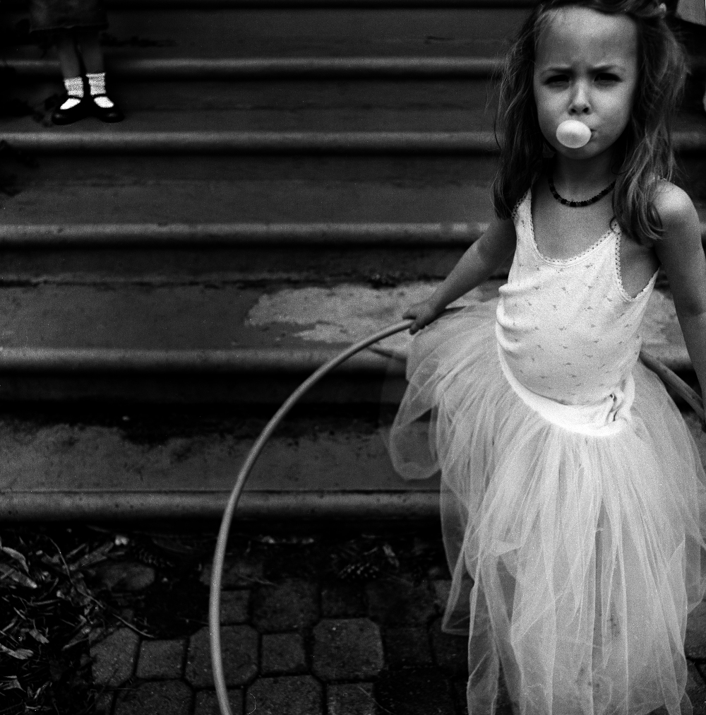 Black and white photograph of a young girl in a white dress sitting on steps while blowing a bubble gum bubble. In the upper left corner, another child's feet wearing white socks and dark shoes are visible on the steps above. A garden hose lies across the lower portion of the composition.