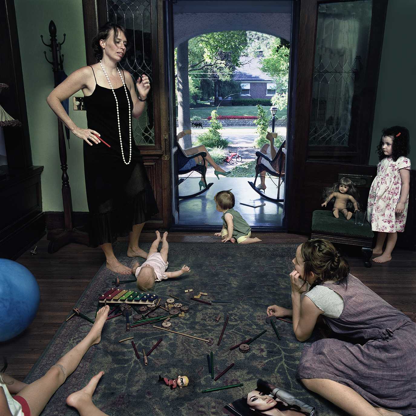 A woman in a black slip dress and long pearl necklace supervises a playroom where several children sit and lie on a gray carpet scattered with toys and dolls. Bright green walls frame the scene, with French doors opening to a sunlit garden beyond, while toys and a blue exercise ball are visible at the edges of the composition.