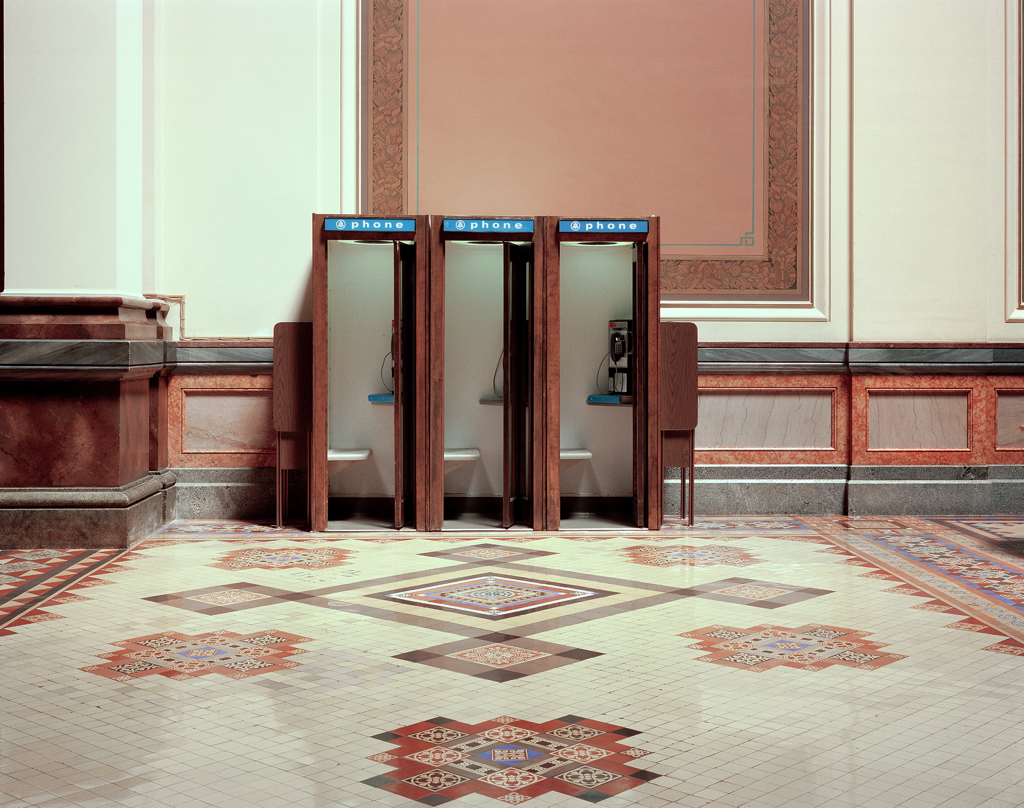 Three wooden phone booths with blue signage stand against a cream and terracotta striped wall in an ornate interior hallway. The floor features an intricate geometric tile pattern in cream, rust, and burgundy tones with symmetrical decorative motifs.