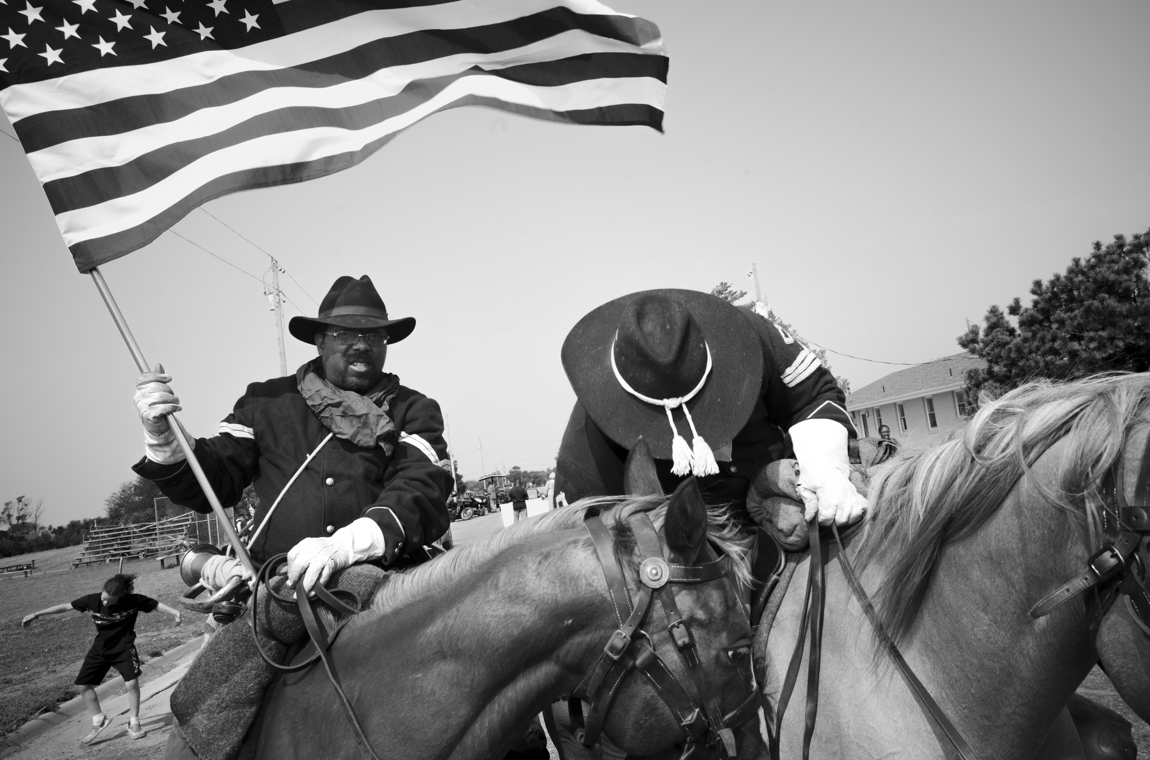 Black and white photograph of two figures on horseback wearing wide-brimmed hats and formal jackets with white gloves, one holding an American flag. A residential area and spectators are visible in the soft-focus background.