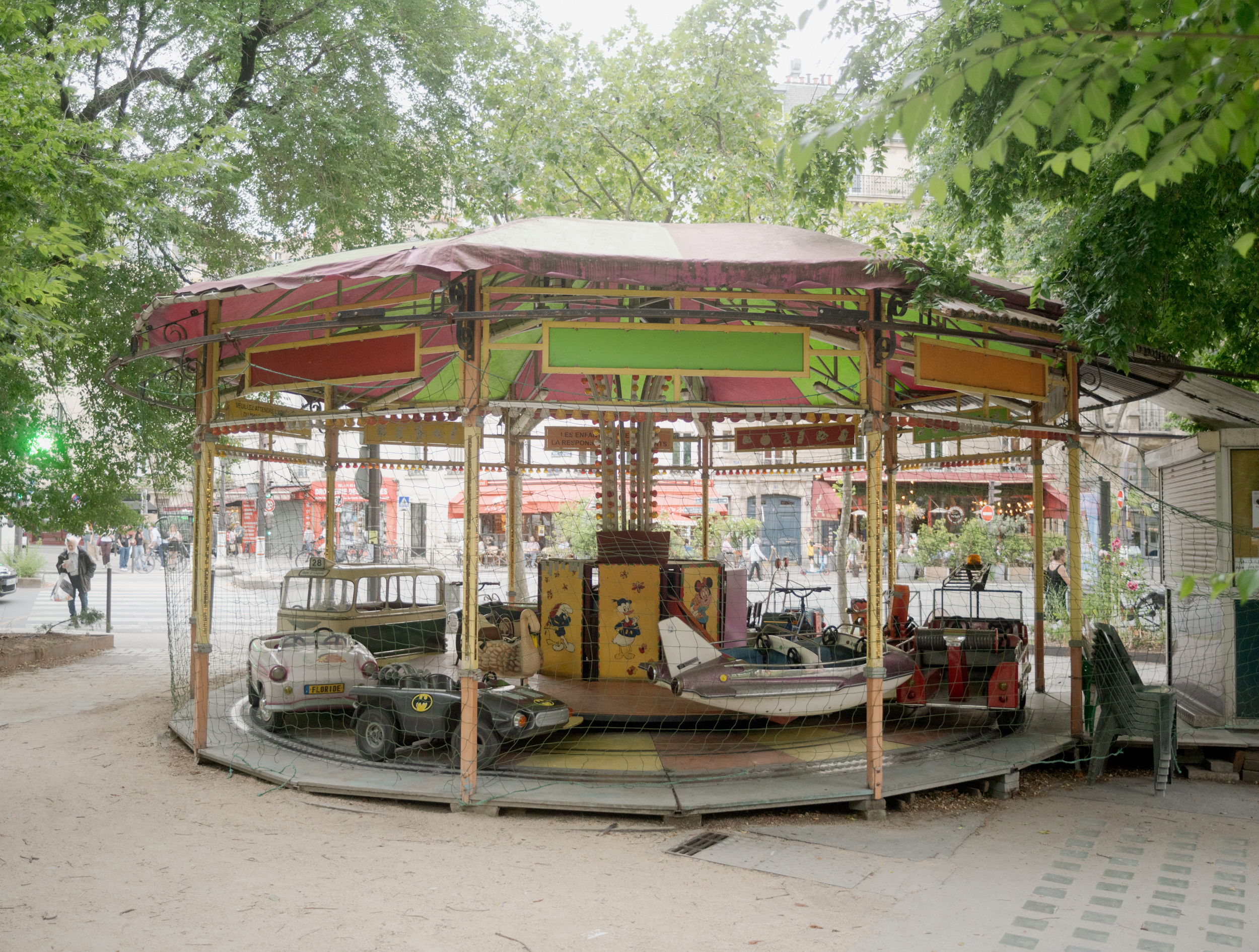 A weathered carousel with a faded red and yellow canopy sits in an urban plaza, its small vehicles painted with comic book characters including Batman. Surrounding trees frame the scene, with brick buildings and pedestrians visible in the soft, overcast light beyond.