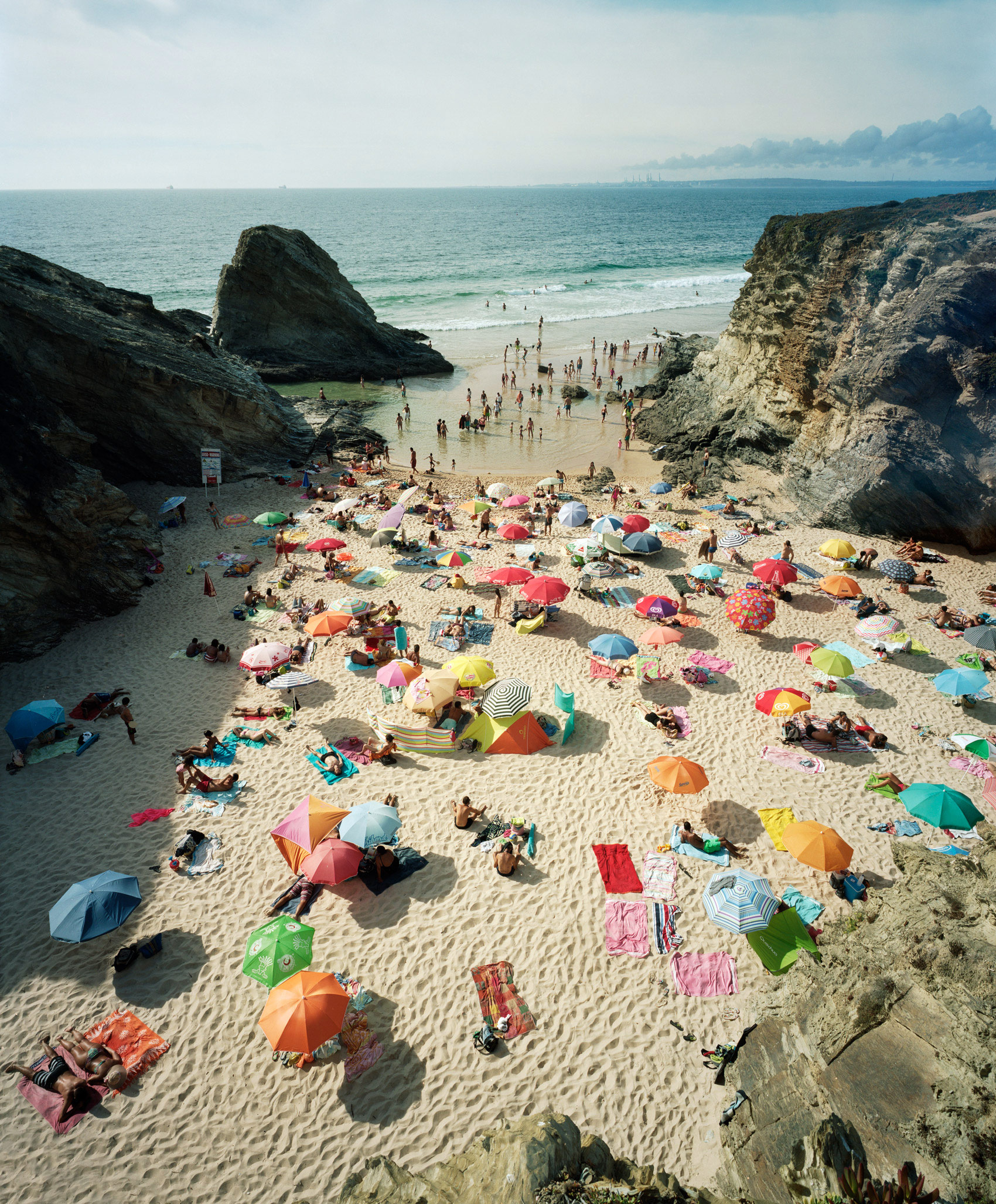 A crowded beach nestled between dramatic rocky cliffs, dotted with numerous colorful umbrellas in red, pink, orange, yellow, blue, and green. Beachgoers occupy the sandy shore and shallow water, their bright parasols creating a vibrant patchwork pattern across the landscape, with the calm sea and distant horizon visible beyond.