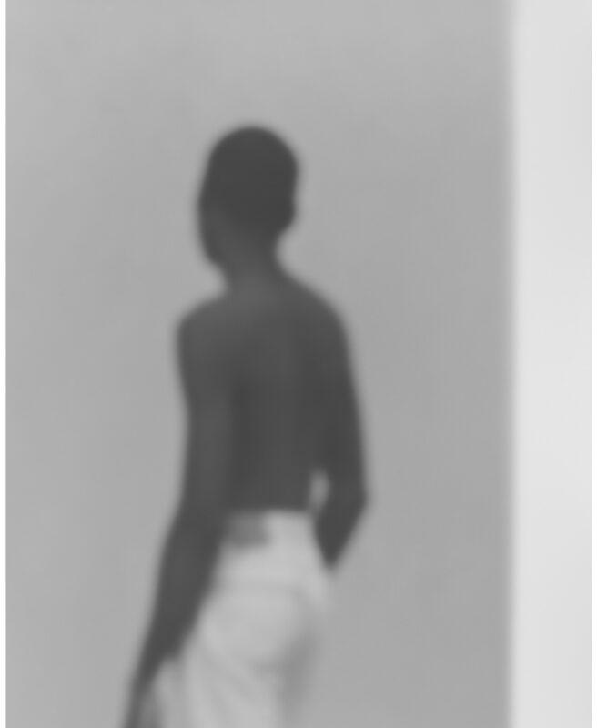 Blurred silhouette of a shirtless figure standing against a light gray wall, rendered in soft black and white tones. The abstracted form and ethereal quality suggest movement or a fleeting moment captured through deliberate focus techniques.
