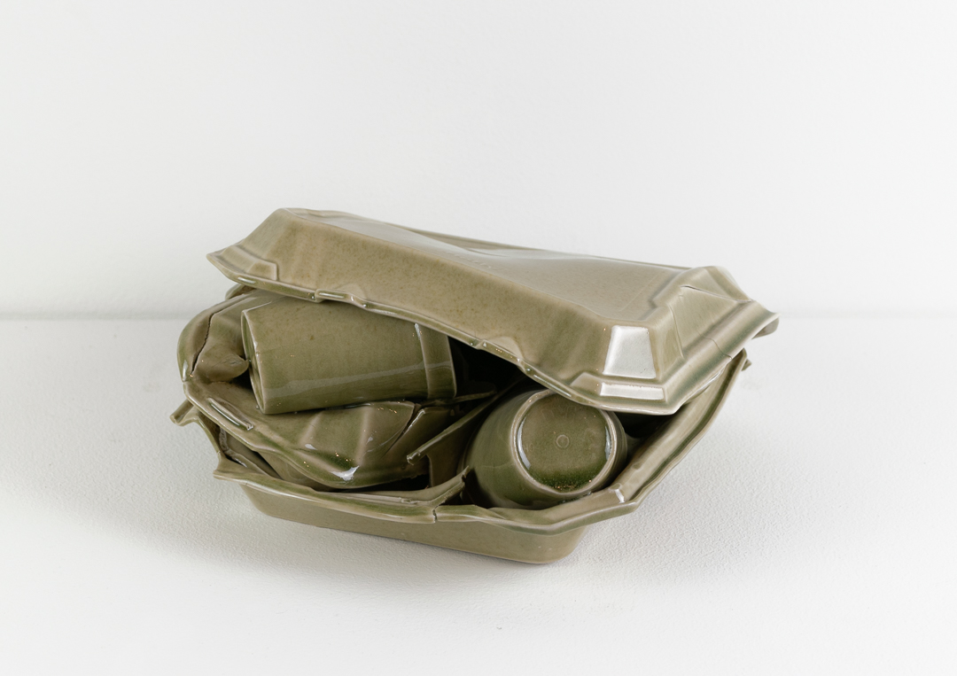 A pale celadon-glazed porcelain sculpture depicting an open takeout container with crumpled paper napkins and two cups nestled inside, rendered in refined ceramic material that mimics the appearance of disposable styrofoam packaging.