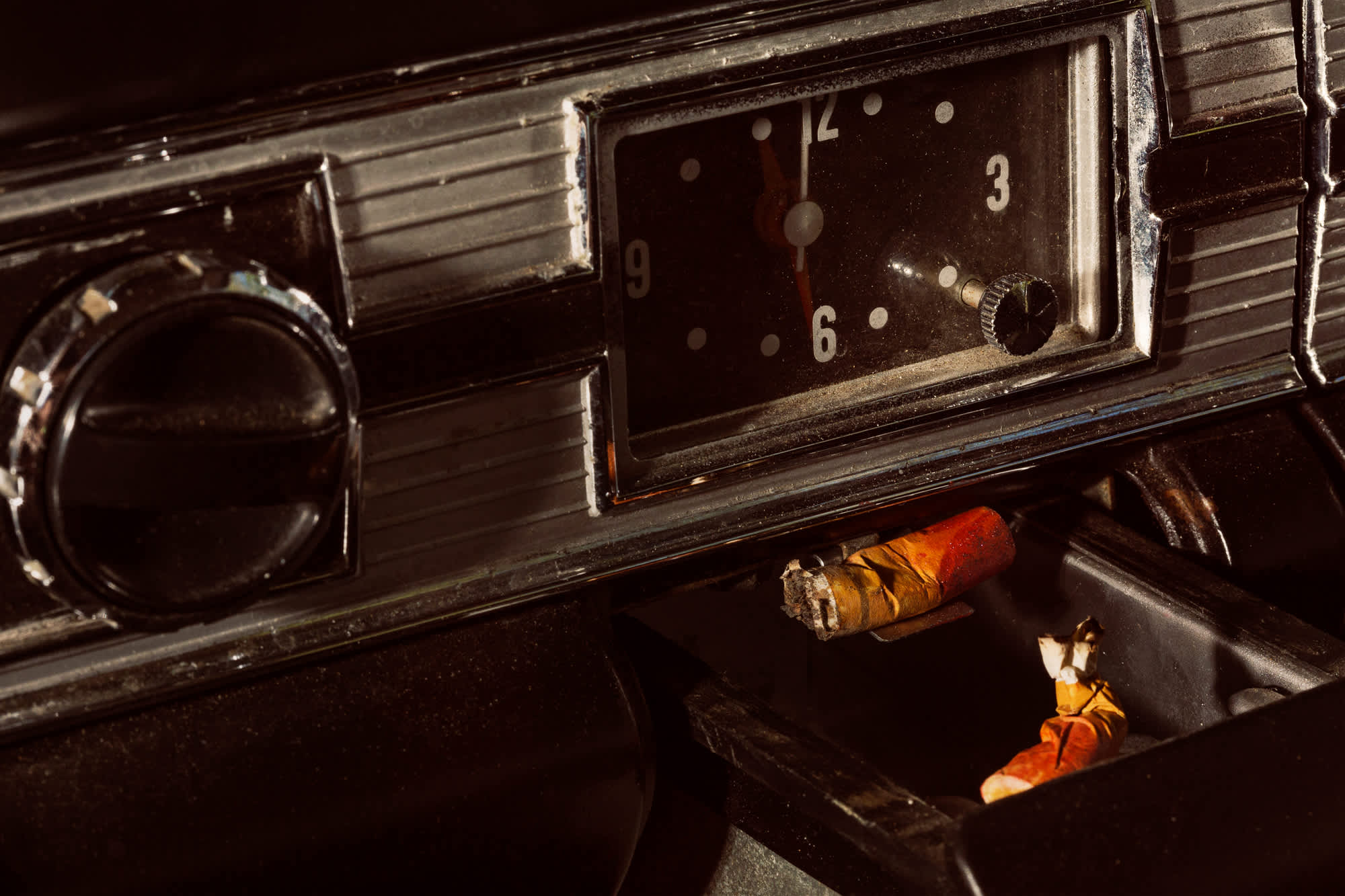 Vintage car dashboard shot from above in dramatic lighting, featuring a dark steering wheel on the left, an analog clock showing approximately 3:00, and a wooden cigar resting on the dashboard. Warm orange and amber tones of the cigar contrast sharply against the black interior and metallic dashboard trim.