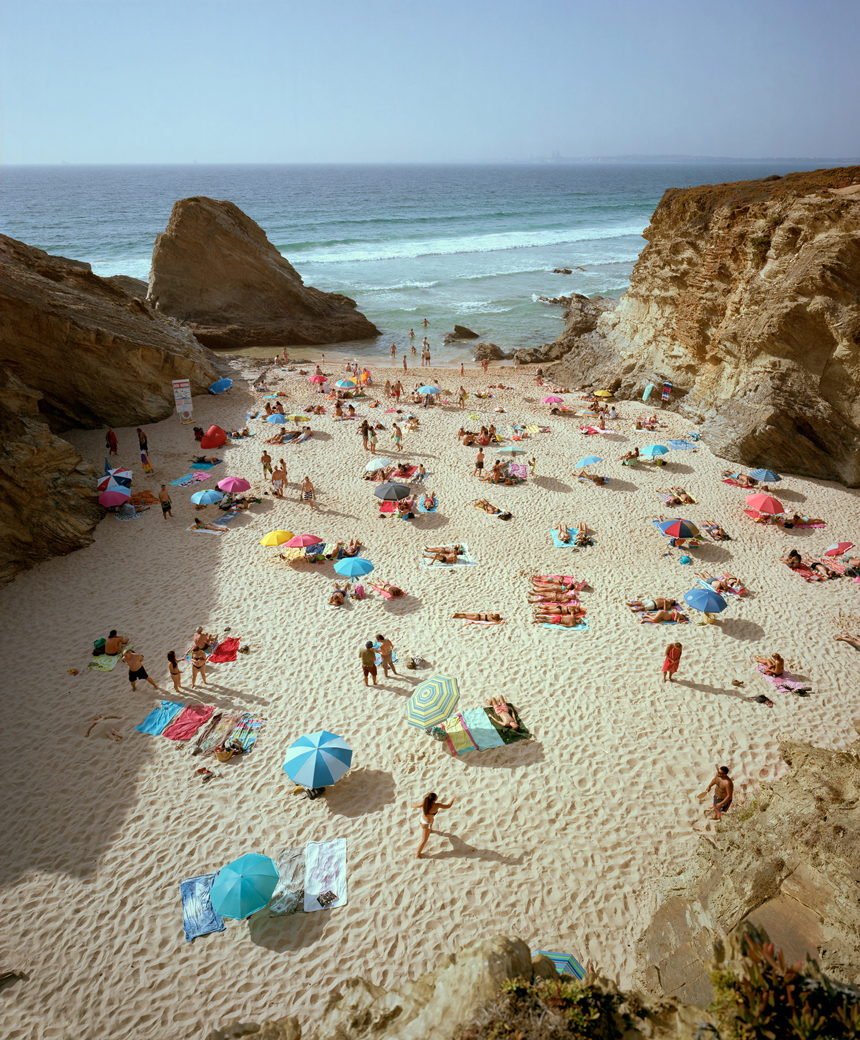 Aerial view of a crowded beach nestled between towering ochre-colored cliff formations, with numerous beachgoers scattered across pale sand beneath colorful umbrellas in shades of blue, pink, and yellow. Turquoise waves and rocky outcroppings frame the scene, while the vast horizon extends across a clear sky.