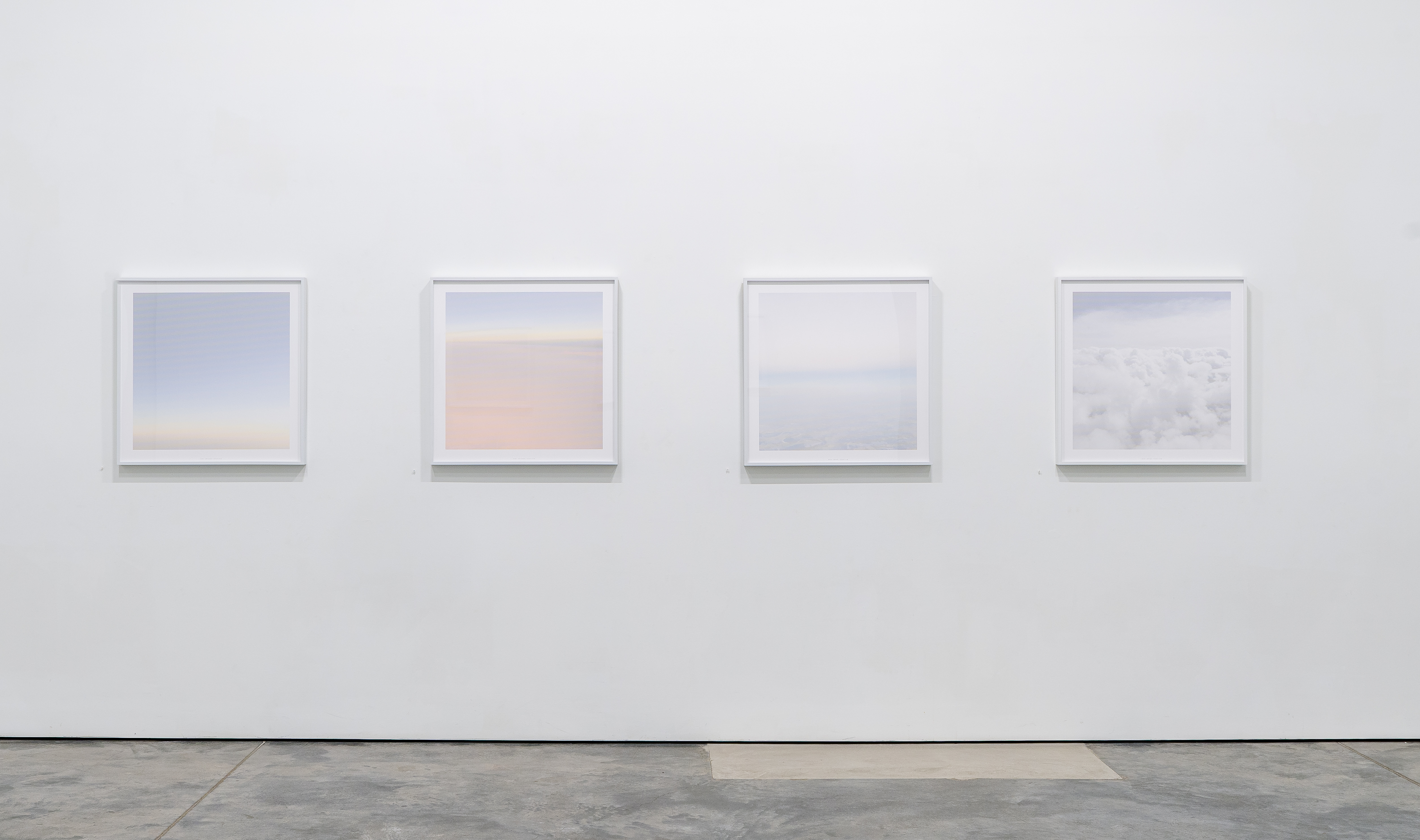 Four framed square photographs mounted in a row on a white gallery wall, each displaying soft, minimalist aerial views of sky, clouds, and horizon in pale blues, pinks, and whites rendered in a dotted halftone technique.