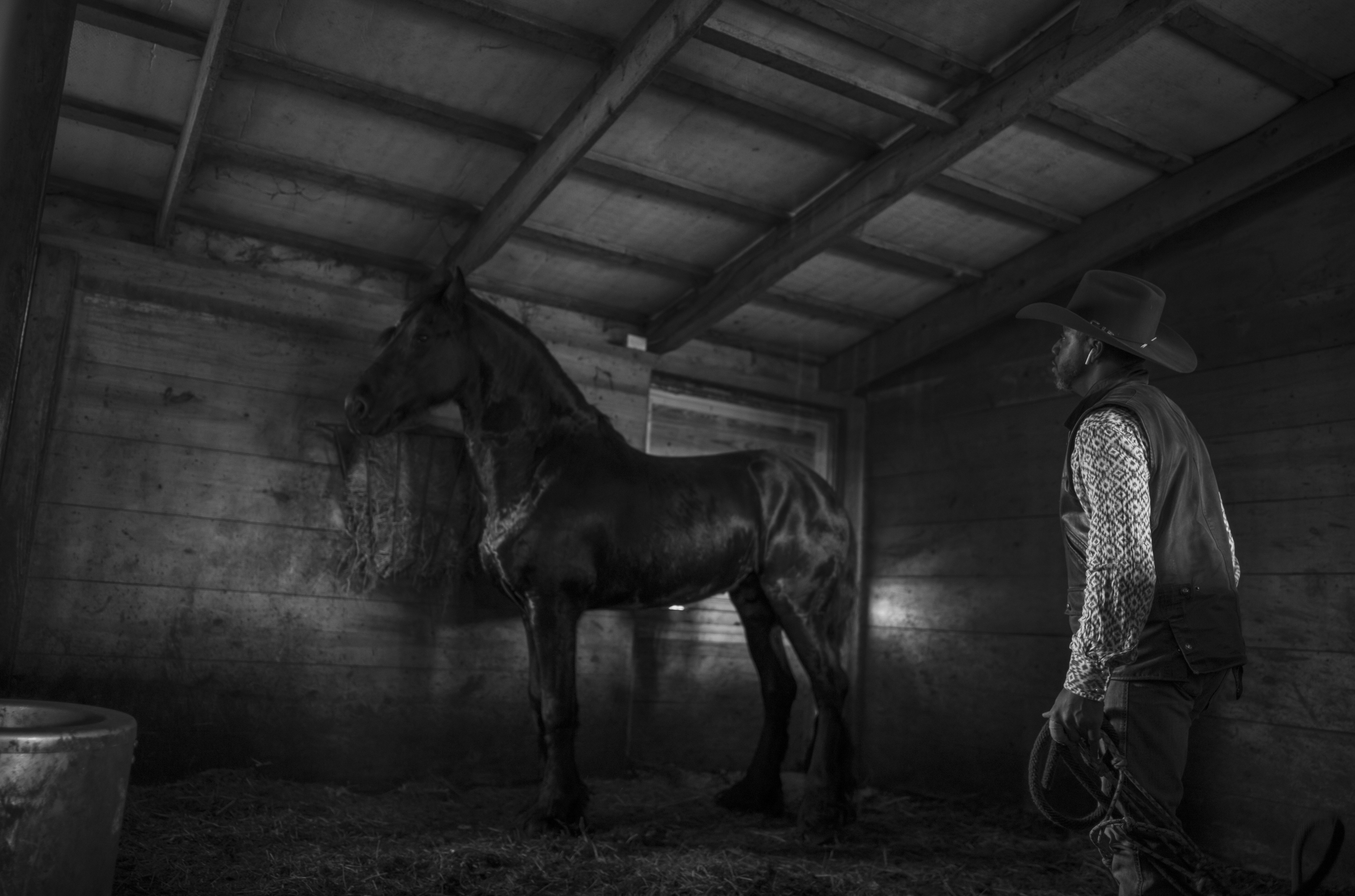 Black and white photograph of a cowboy in a wide-brimmed hat and patterned vest standing beside a dark horse inside a wooden barn structure, shot in high contrast with dramatic lighting that emphasizes the textures of weathered wood beams and earthen floor.