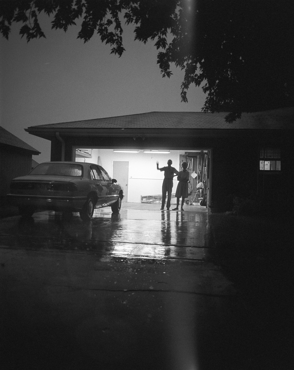 Black and white photograph of a wet garage interior at night, with fluorescent lighting illuminating two silhouetted figures standing in the doorway. A dark sedan is parked on the left side of the reflective wet floor, and hanging storage items are visible in the background. Tree branches with foliage hang from above, partially framing the scene.