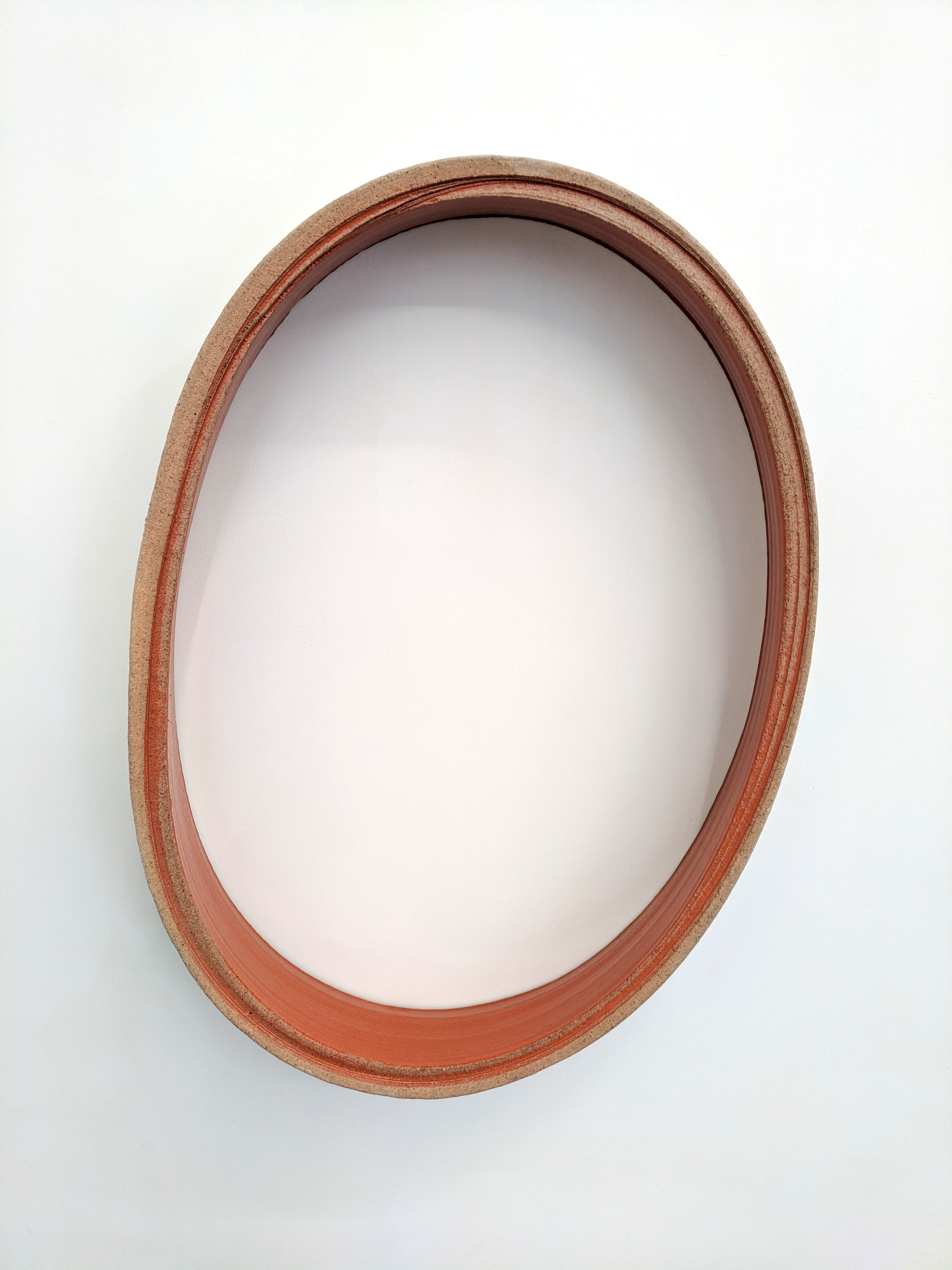 An oval ceramic frame with a terracotta-red interior band and weathered tan outer edge, mounted vertically against a white wall with an empty white center.