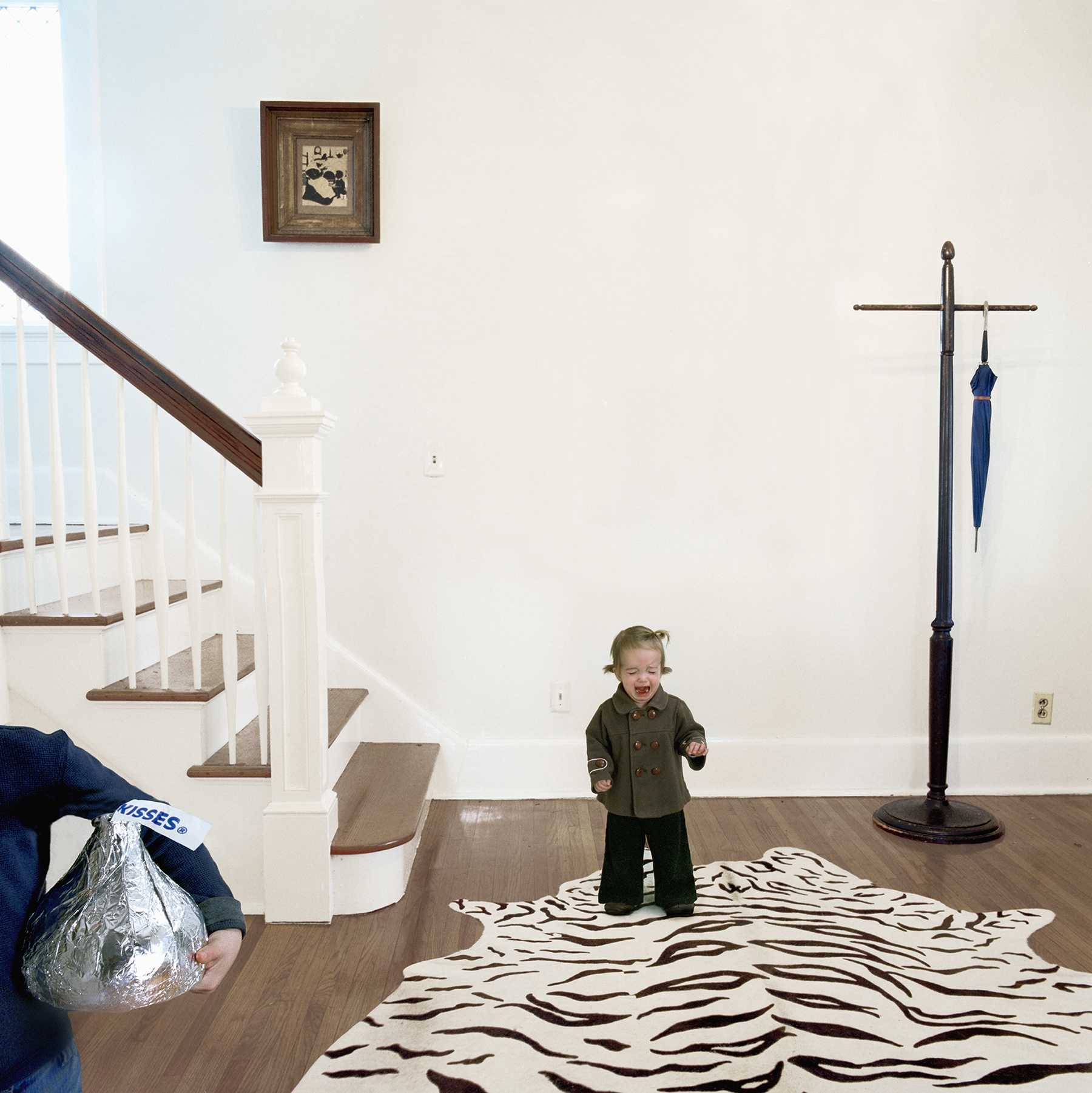 A young child in an olive-green jacket stands on a zebra-striped rug in a bright, minimalist interior space, appearing to laugh or call out. The composition includes a white staircase with wooden treads on the left, a tall dark wooden cross with a blue sword on the right wall, and framed artwork above, while an adult's legs and a silver shopping bag are visible in the lower left corner.