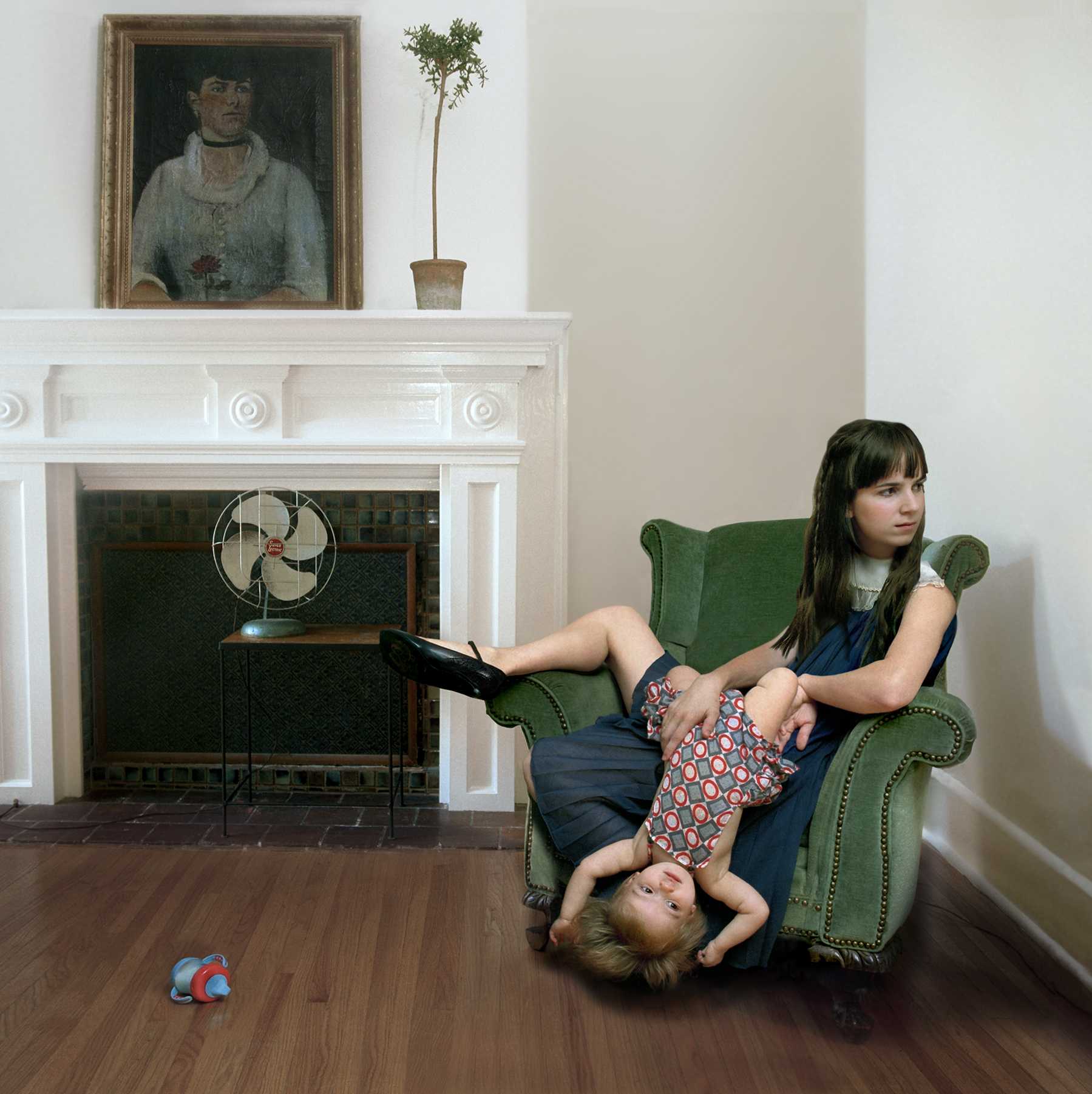 A woman and young child sit together on a green upholstered armchair in a bright, classically decorated interior. The woman holds a book while the child reclines across her lap with legs extended. Behind them, a white fireplace mantel displays a framed portrait and a potted plant, with a vintage fan visible in the fireplace opening. Warm wood flooring and neutral walls frame the domestic scene.