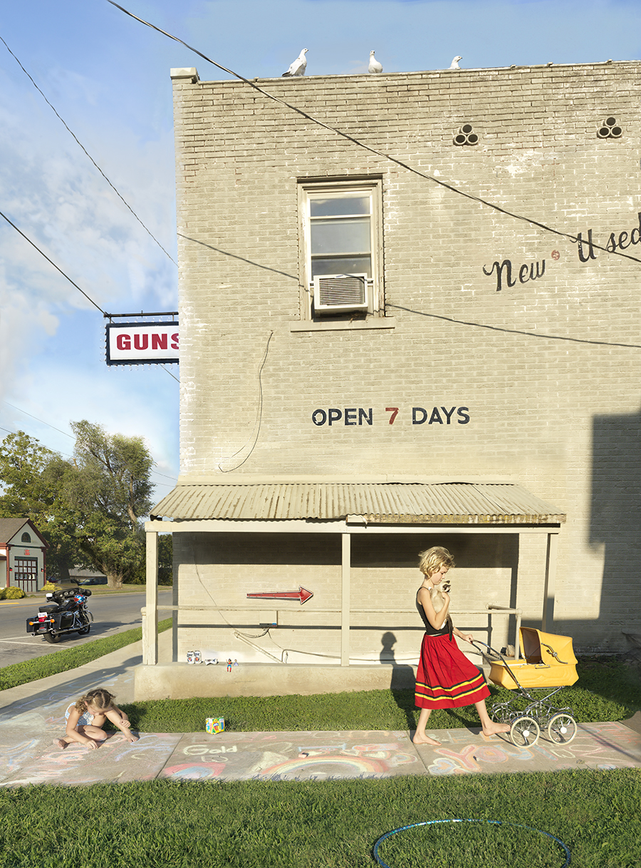 A staged tableau depicts children playing in front of a brick building with "GUNS" signage and "OPEN 7 DAYS" painted on its facade. The scene juxtaposes childhood innocence—chalk drawings, toys, and play—against the austere commercial architecture and weaponry commerce suggested by the storefront.