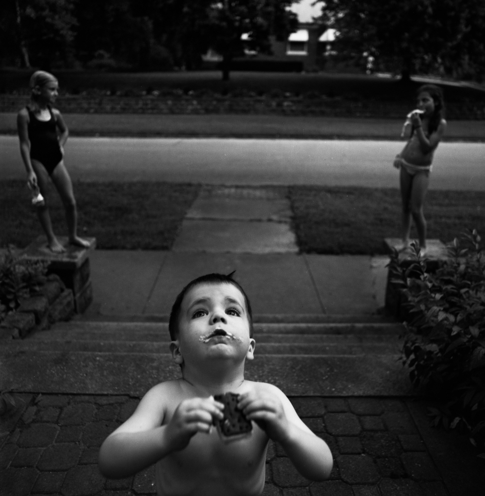 Black and white photograph of a shirtless young child in the foreground looking upward, holding what appears to be a melting treat, set in a residential outdoor space with a paved area and two girls standing in the background near a pool.