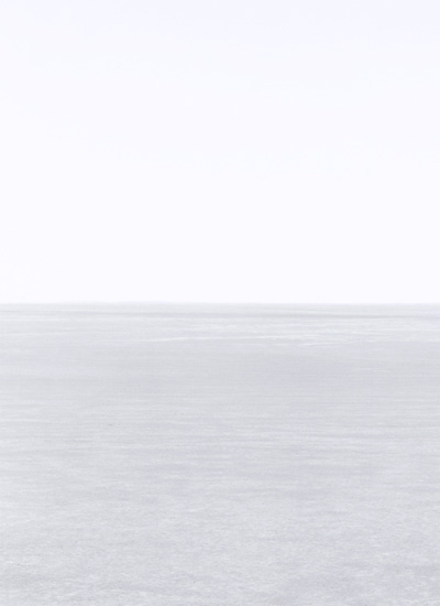 Minimalist landscape divided horizontally into pale sky above and textured white-grey ground below, with a subtle horizon line creating a stark, contemplative composition in soft, muted tones.