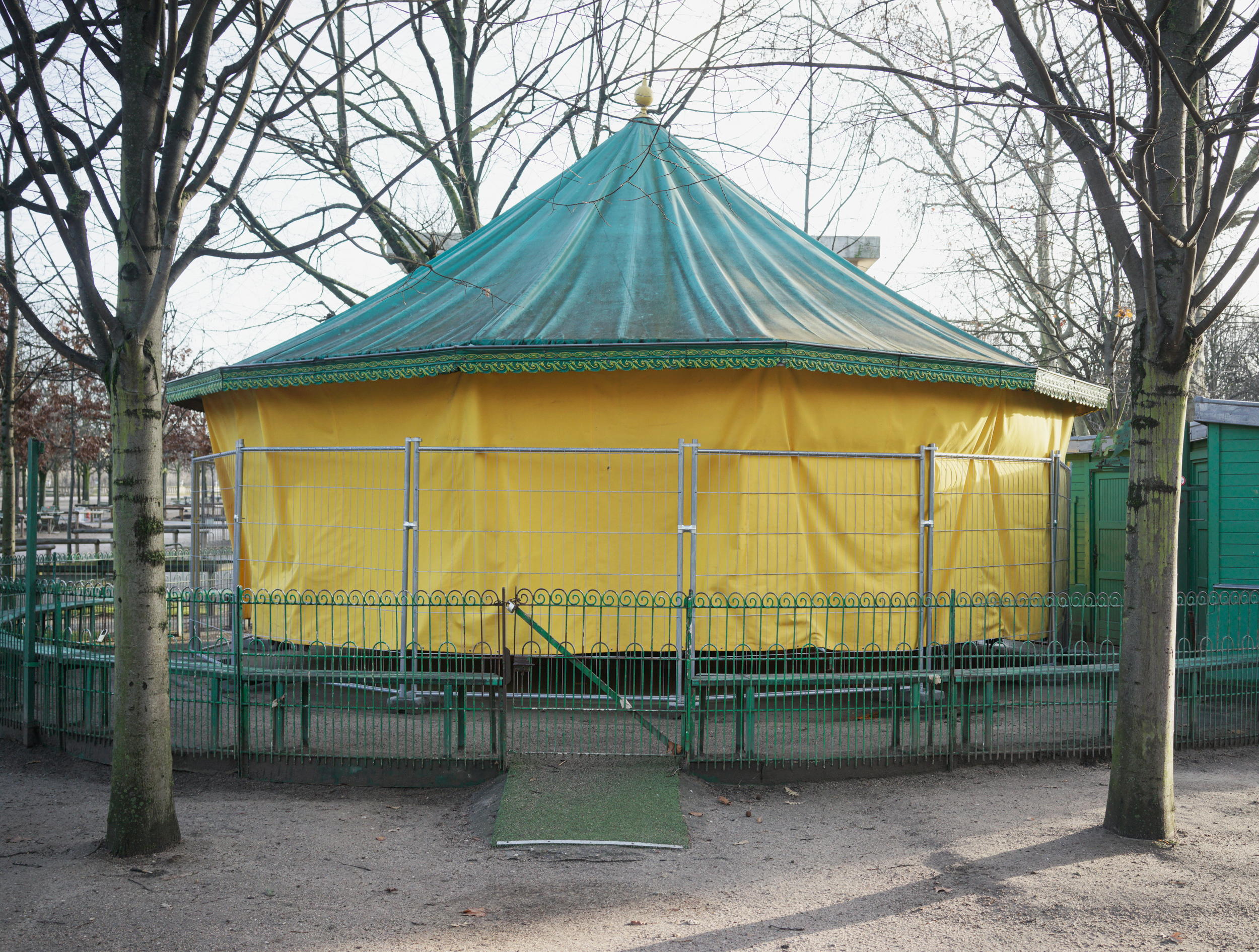 A circular bandstand with a turquoise conical roof and yellow fabric walls stands in a park, framed by bare trees and surrounded by green metal fencing. The structure sits on a gravel ground with ornamental railings, creating a striking contrast of bright colors against the muted winter landscape.