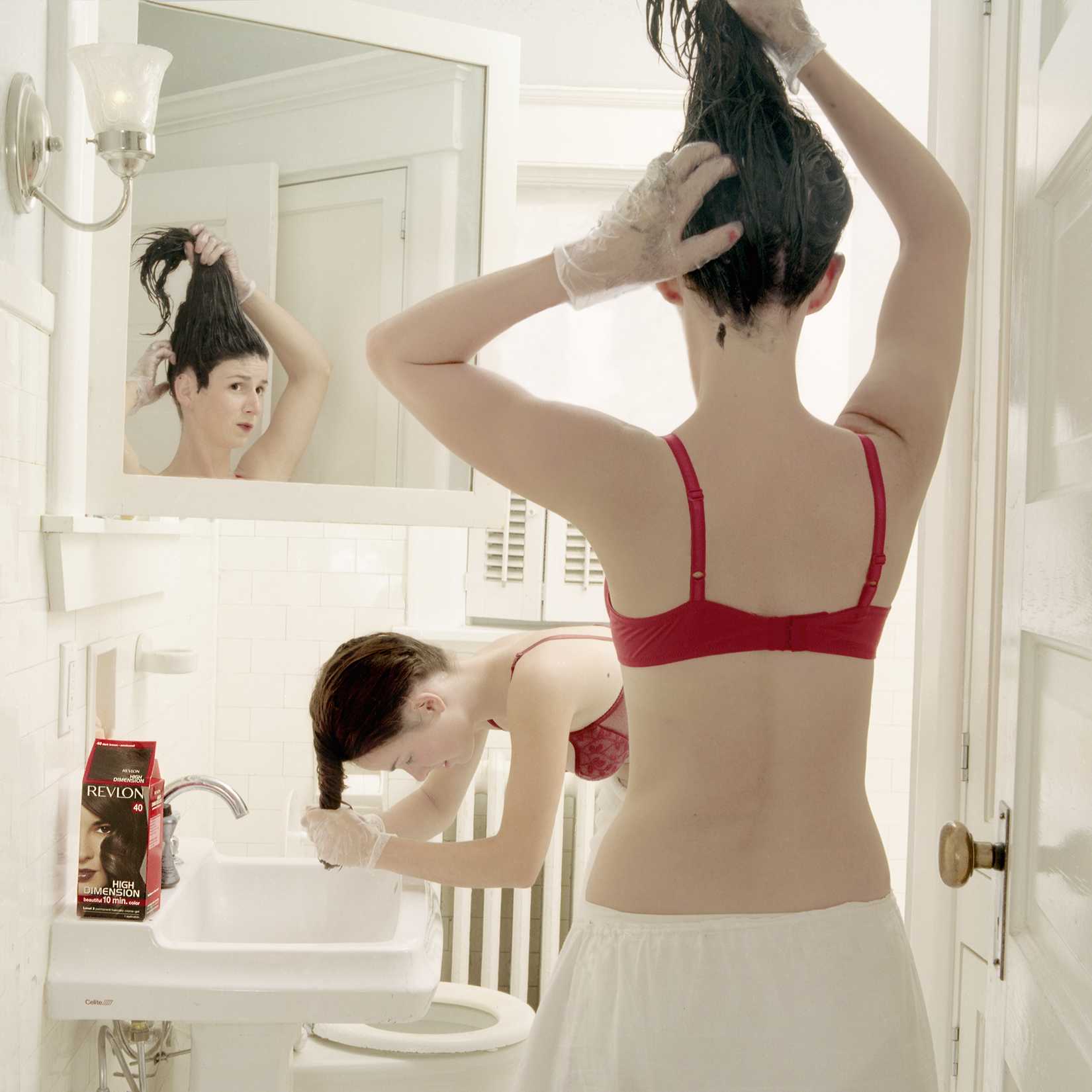 A woman in a red bra and white skirt stands at a bathroom sink, viewed from behind as she lifts her dark hair above her head. Her reflection appears in the large mirror, showing her face in profile, while a bottle of Revlon hair product sits on the sink counter below.