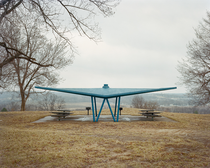 A large teal-blue sculptural shelter with an expansive wing-like roof structure stands in a sparse winter landscape. Bare trees frame the composition on either side, with picnic tables positioned beneath the canopy and rolling hills visible in the distant background.