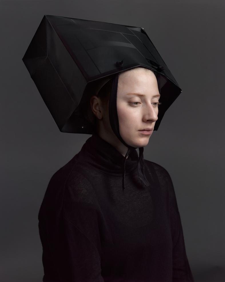 A figure in profile wearing a dark, high-necked garment with a striking geometric black hood that extends outward in angular, box-like planes. The minimalist composition is set against a neutral gray background, emphasizing the sculptural quality of the headpiece and the contemplative downward gaze of the pale-faced subject.