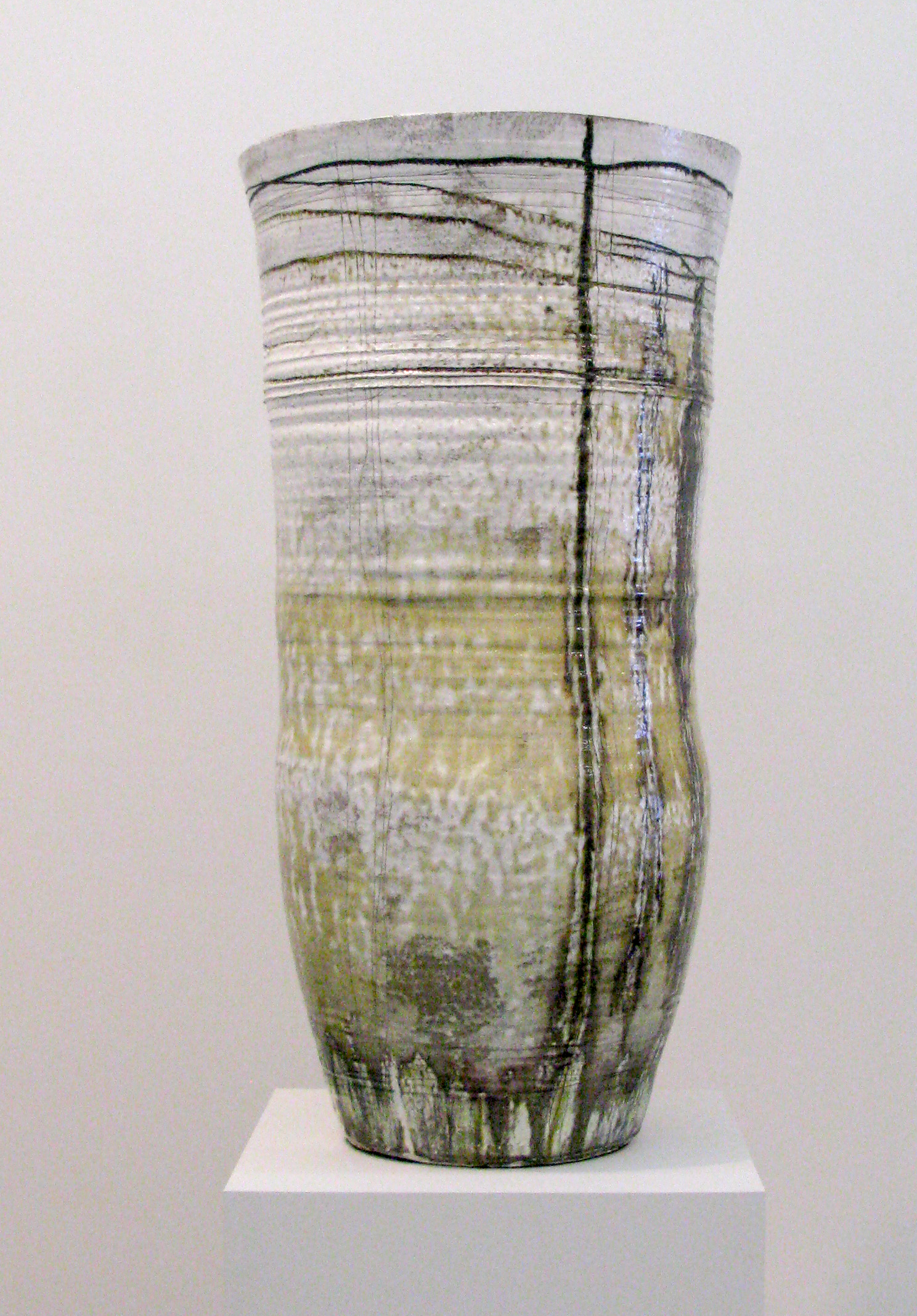 A tall cylindrical stoneware vessel with a matte surface featuring horizontal linear markings and vertical incised lines. The vessel transitions in color from pale gray-white at the rim to muted green and ochre tones toward the base, with darker accretions suggesting natural weathering or firing effects.