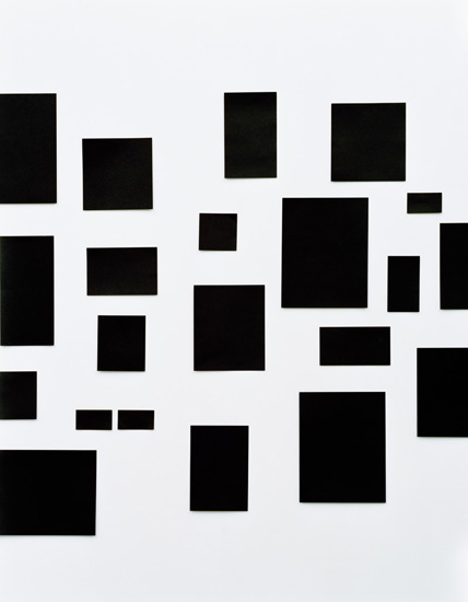 Abstract composition of scattered black rectangular forms of varying sizes arranged across a light gray background, creating a fragmented, geometric spatial arrangement.