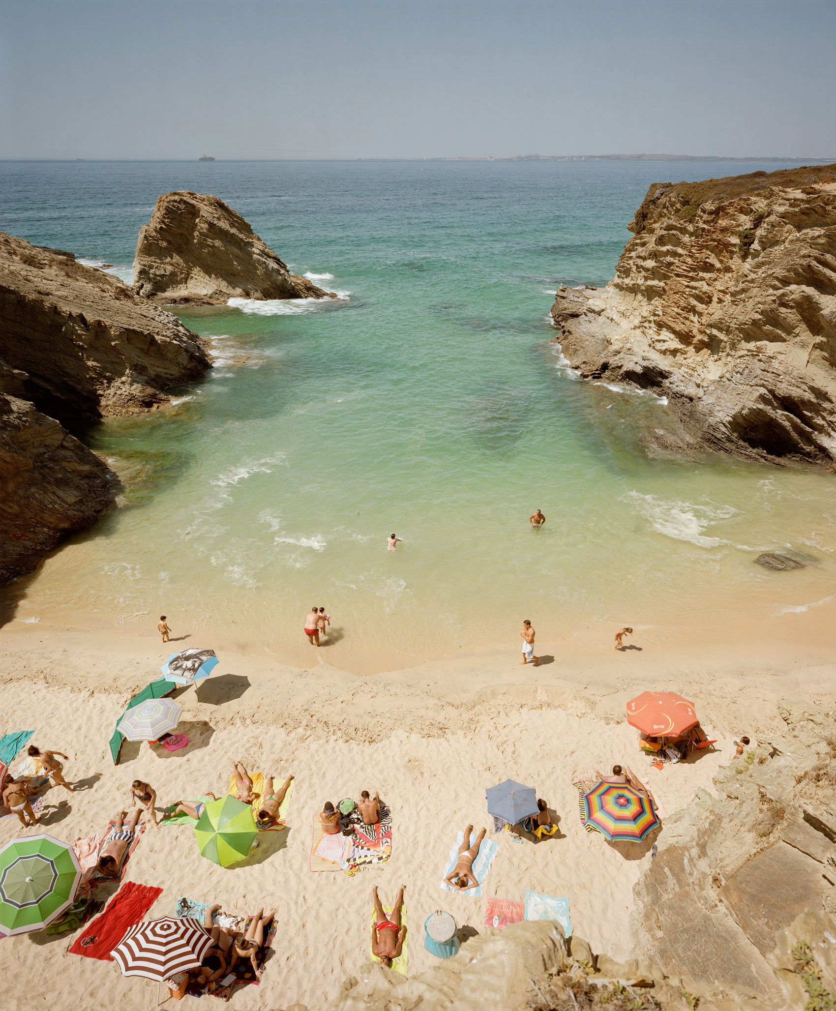 Aerial view of a sheltered beach cove framed by dramatic limestone cliffs, with turquoise water and sandy shore dotted with colorful beach umbrellas, towels, and sunbathers scattered across the sand.