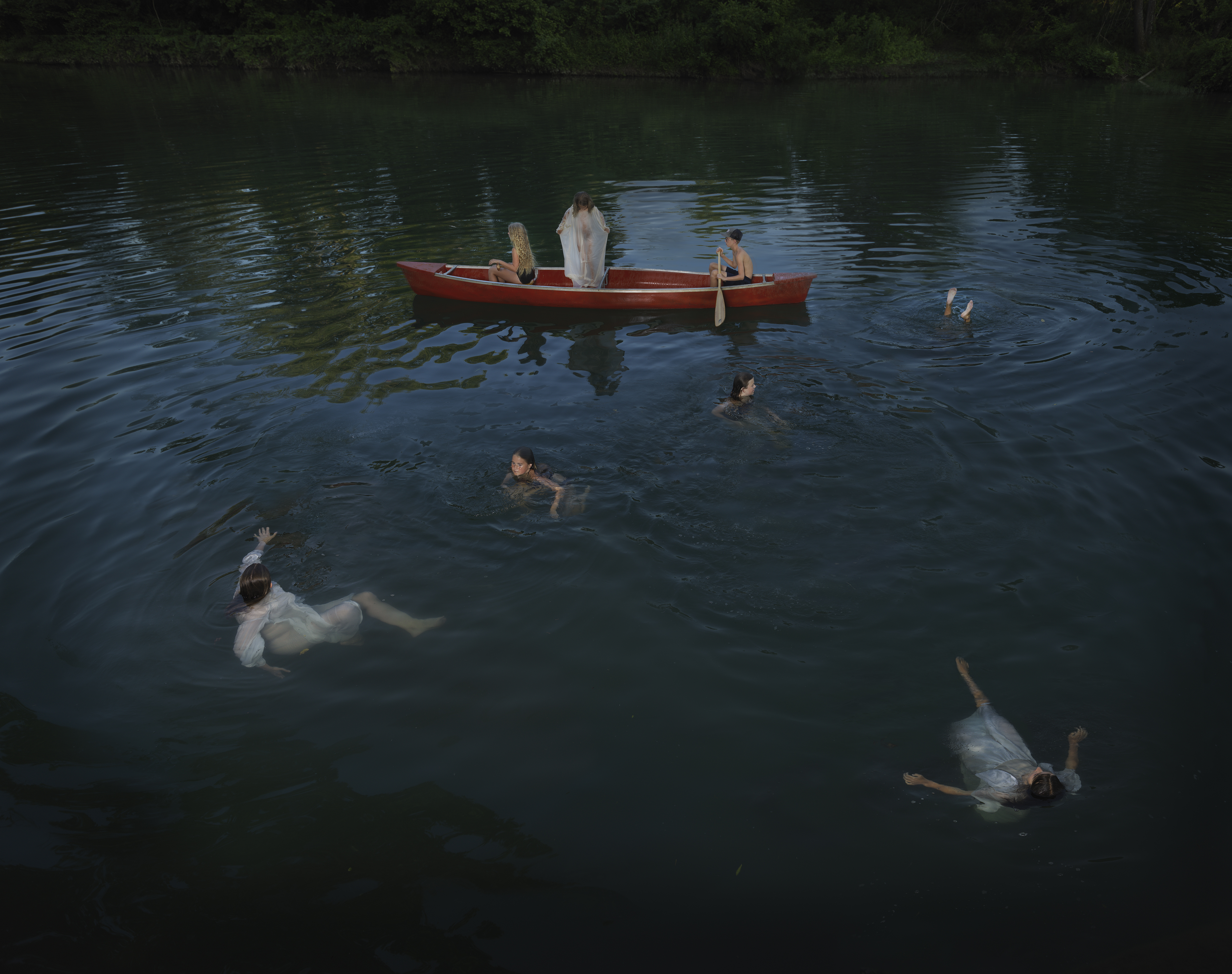 Several figures in white clothing float and swim in dark water surrounded by dense forest, while a red rowboat with two occupants sits anchored in the center of the composition. The cool, moody lighting creates a dreamlike atmosphere across the still water's surface.